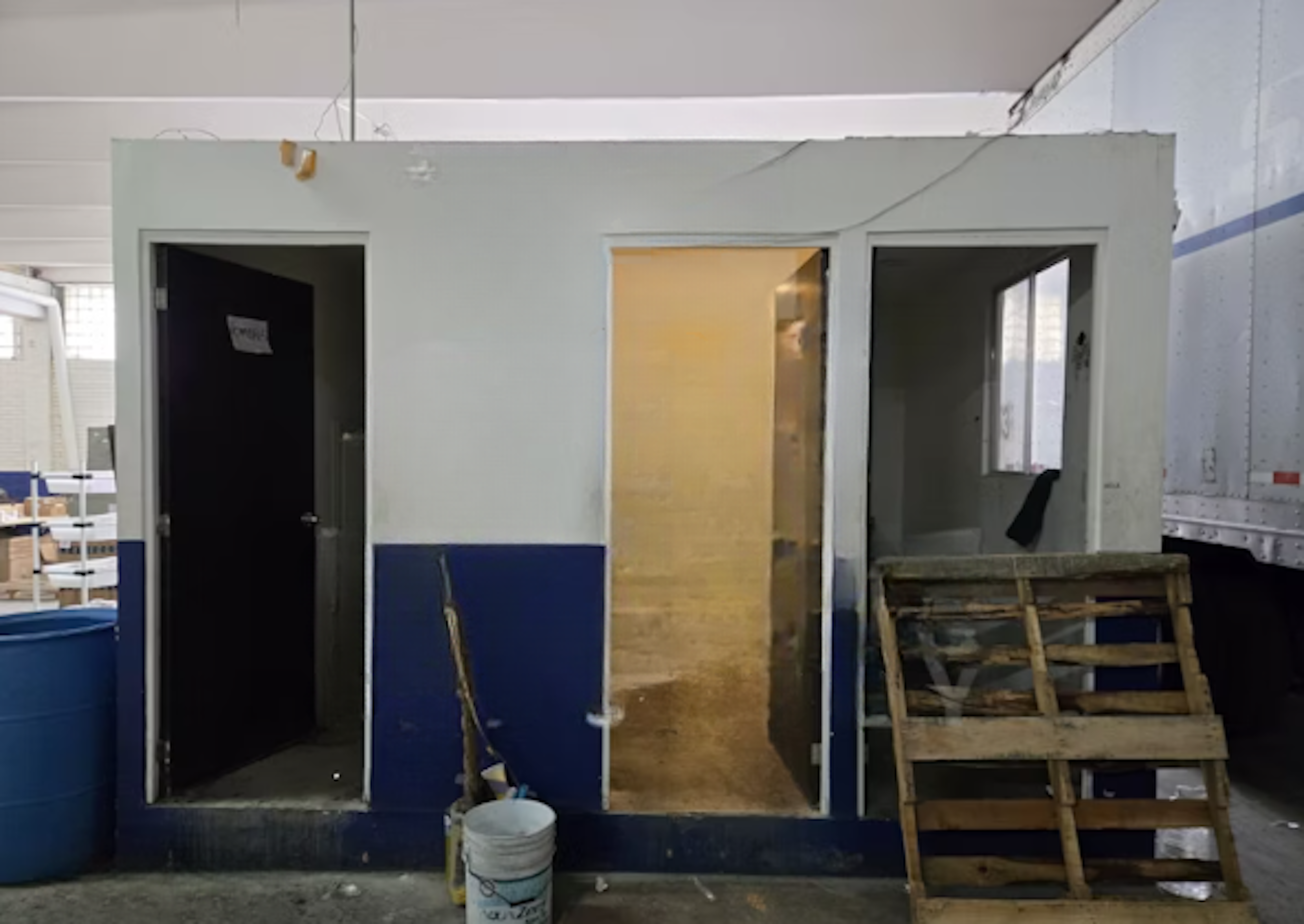 La imagen muestra una construcción en progreso de una pequeña estructura modular con tres puertas. Parece ser un área de oficinas o vestidores temporales en un sitio de construcción. Las paredes están pintadas de blanco y azul, con una puerta central iluminada. Hay elementos de construcción visibles como un cubo, una escalera de madera y algunos escombros, indicando que el trabajo aún está en curso.