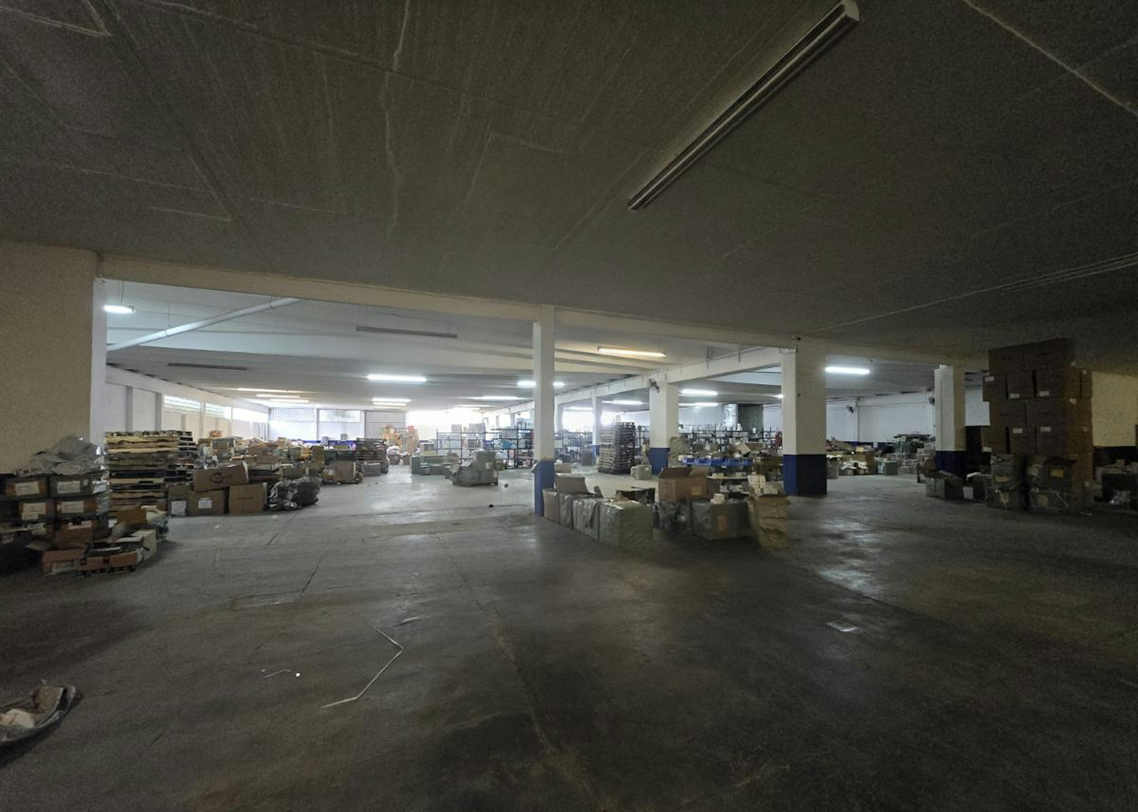 La imagen muestra un amplio almacén o centro de distribución logística industrial. Se observa un espacio interior extenso con columnas, iluminación fluorescente y estanterías. Hay cajas y pallets dispersos por el suelo. En el techo se proyecta un logo que representa la industria logística, con iconos de camión, avión y cajas. El texto Strategic Industry Logistic y INDUSTRY se muestra en el suelo, indicando el propósito del espacio.