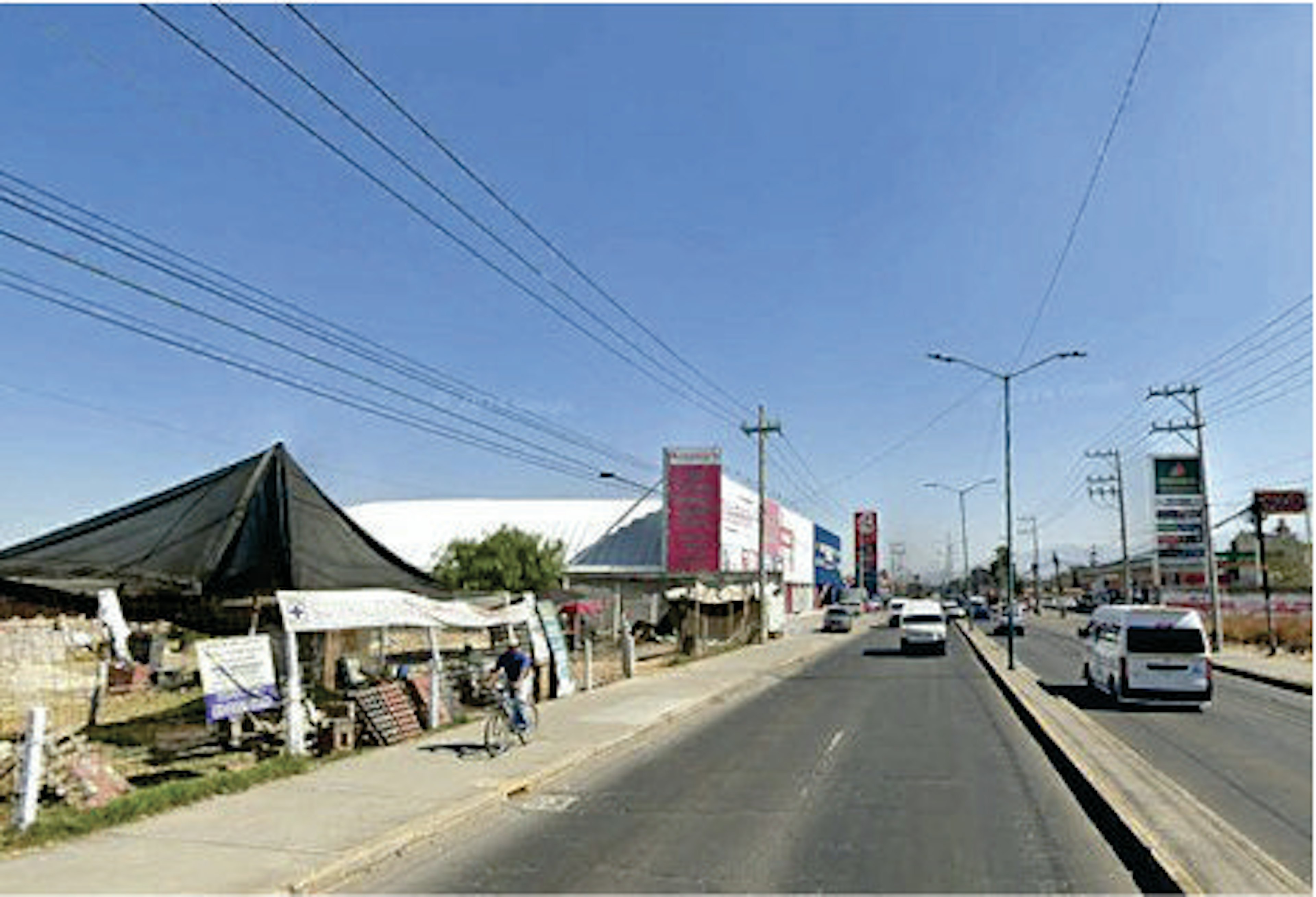 Vista de una calle comercial con varios carteles y establecimientos a los lados. Hay vehículos en la carretera y cables de electricidad visibles en el cielo despejado.