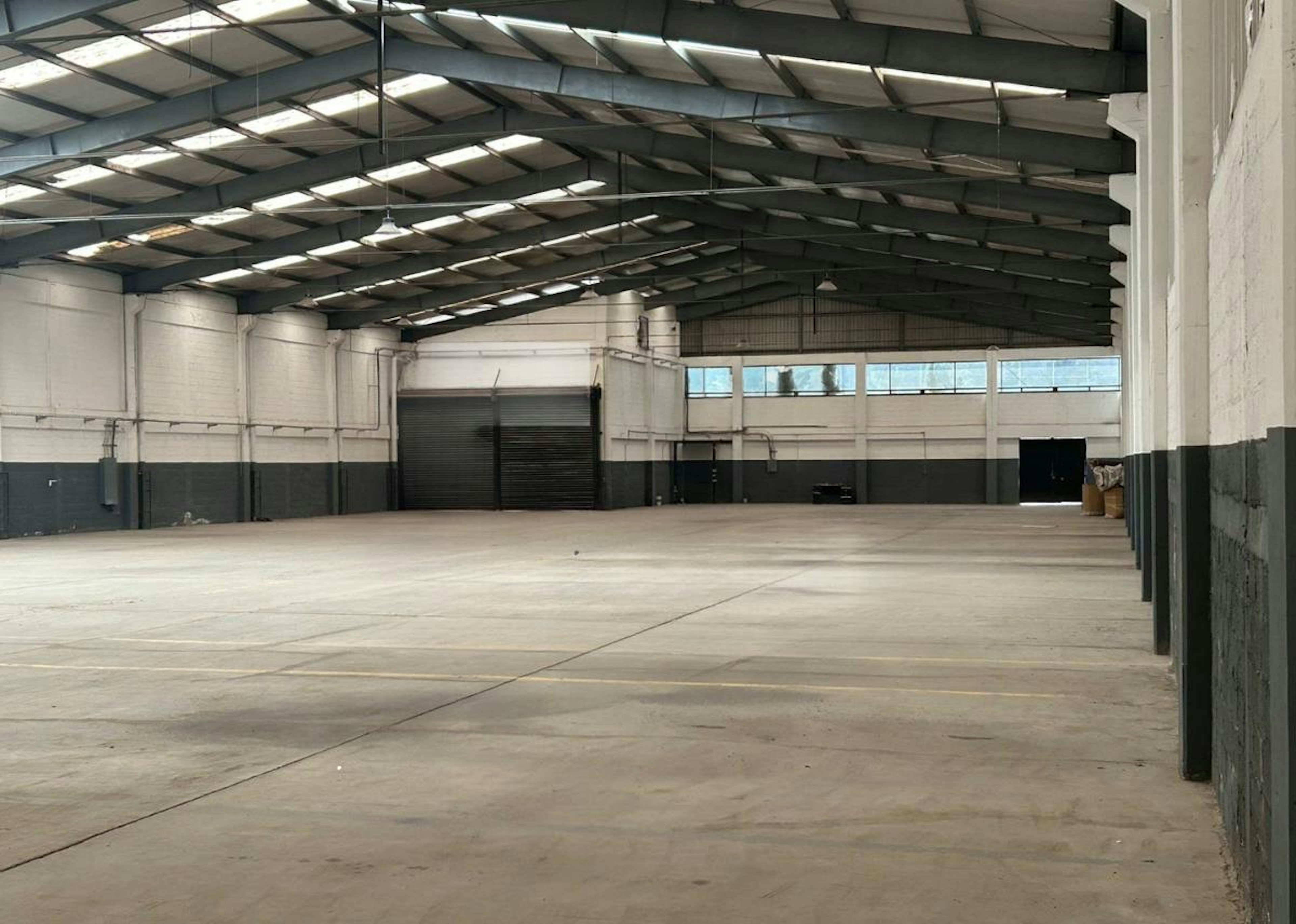La imagen muestra un amplio espacio industrial interior, probablemente un almacén o nave logística. Tiene un techo alto con vigas expuestas y luces fluorescentes. Las paredes son blancas en la parte superior y grises en la inferior. El piso es de concreto y hay algunas puertas enrollables visibles. En el primer plano se ve un logo transparente que dice INDUSTRY y Strategic Industrial Logistics.