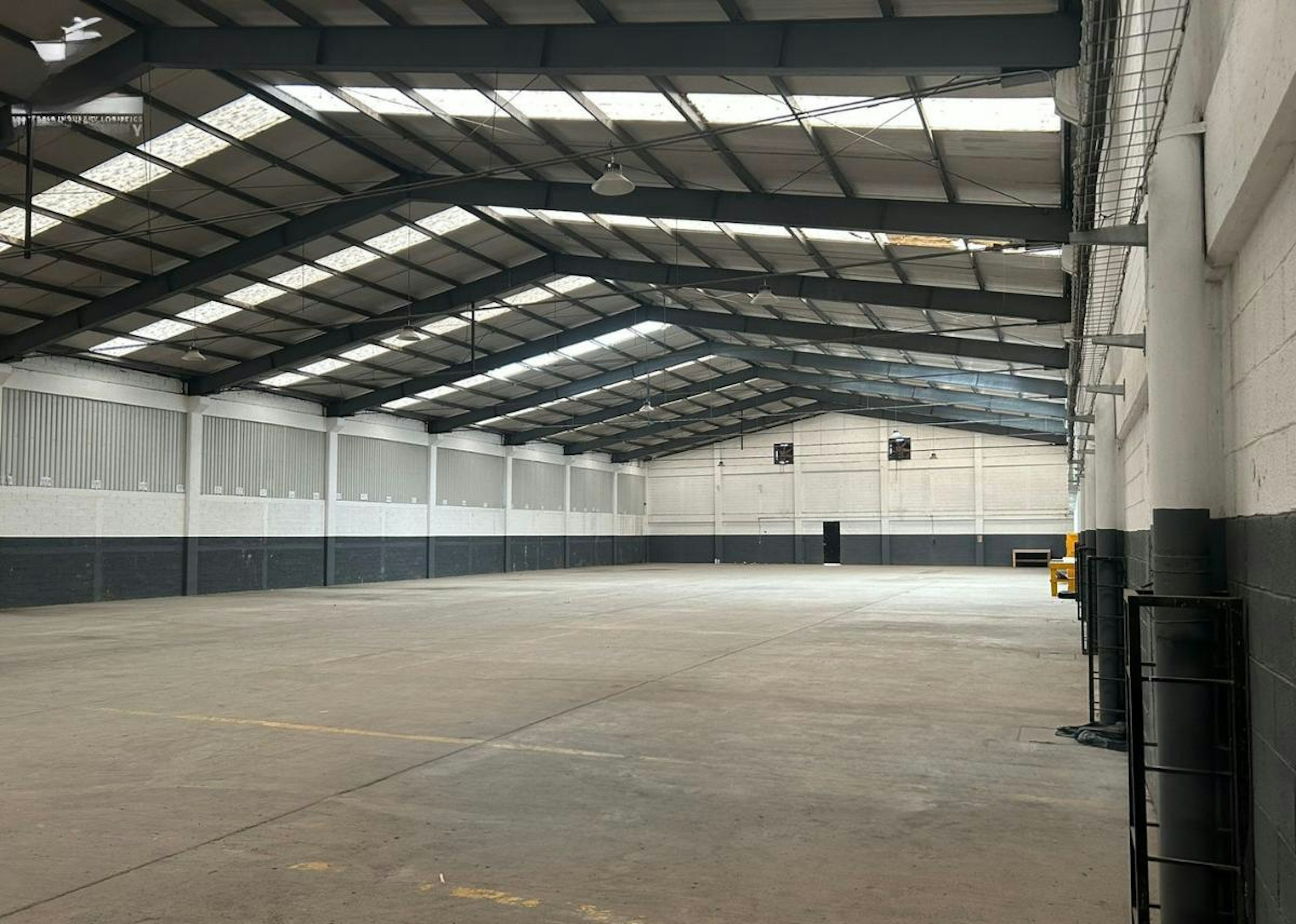 La imagen muestra un amplio almacén o nave industrial vacía con techo alto de estructura metálica y tragaluces. Las paredes son blancas en la parte superior y grises en la inferior. El piso es de concreto y está despejado. Se observan algunos elementos industriales como barriles en un costado. La iluminación natural entra por el techo, creando un espacio luminoso y funcional para uso industrial o logístico.