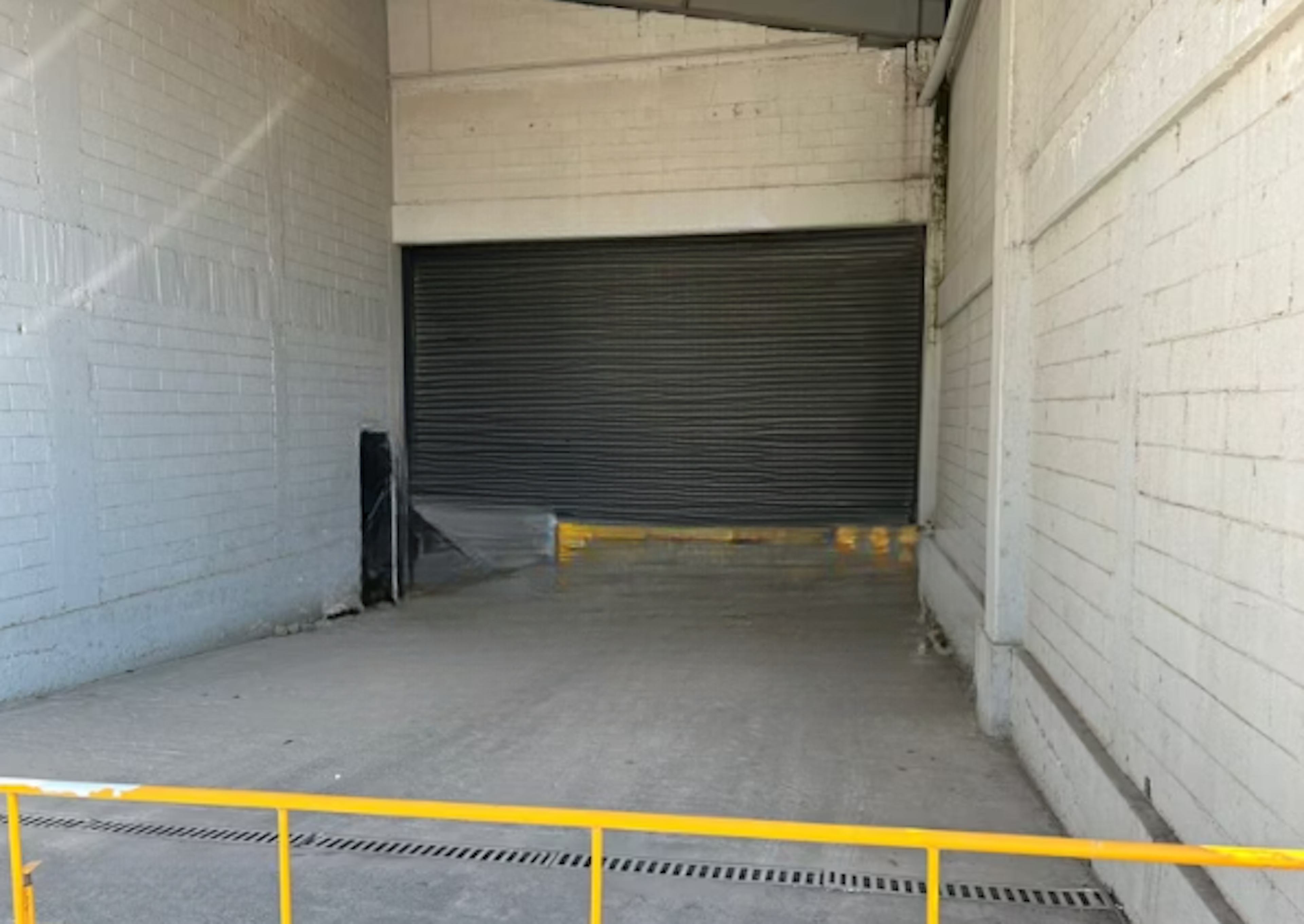 La imagen muestra la entrada a un almacén o garaje industrial. Se ve una puerta de persiana metálica cerrada al fondo, rodeada de paredes de ladrillo pintadas de blanco. El suelo es de concreto y hay una barrera de seguridad amarilla en primer plano. El espacio parece estar en un área de carga o descarga, con un drenaje visible en el suelo. La estética es funcional y utilitaria, típica de espacios industriales o comerciales.