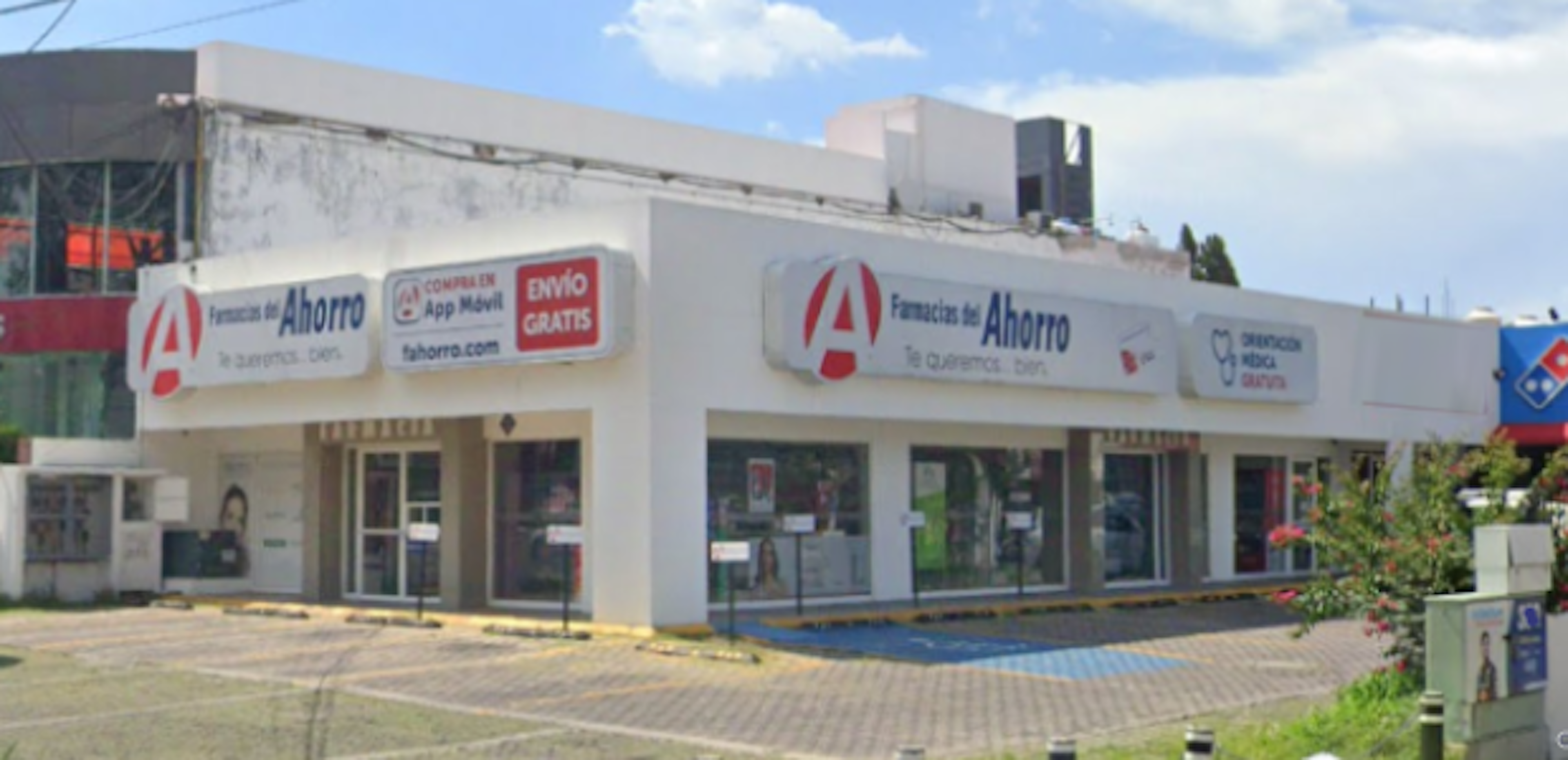 La imagen muestra la fachada de una farmacia llamada Farmacias del Ahorro. Es un edificio comercial de una planta, pintado de blanco con letreros rojos y azules. Tiene amplios ventanales y puertas de vidrio. El local parece estar ubicado en una zona comercial, con un estacionamiento pavimentado al frente. Se observa parte de otro negocio adyacente y vegetación alrededor.