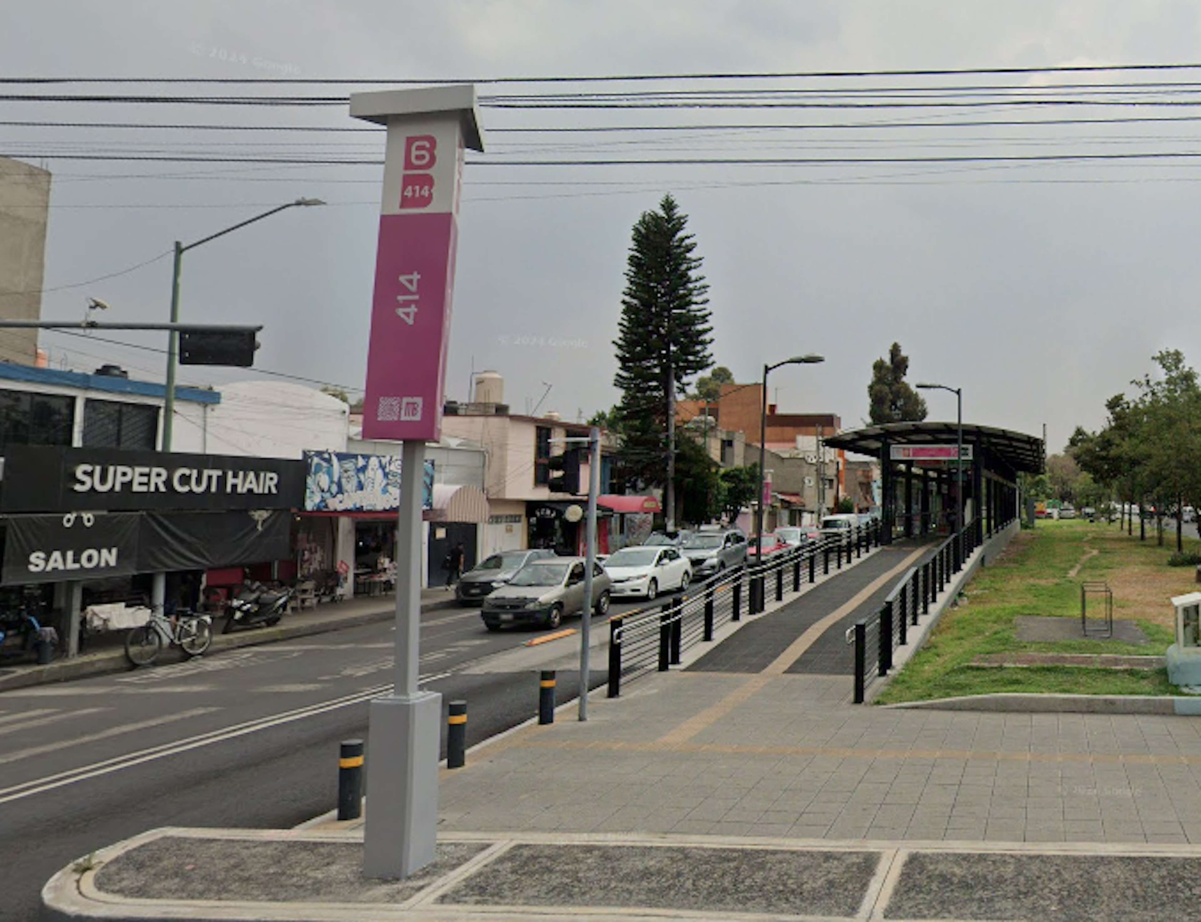 La imagen muestra una calle urbana con una parada de transporte público, identificada por un poste rosa con el número 414. Hay una acera amplia con barandillas, árboles y una estructura cubierta para esperar. Se ven negocios locales como un salón de belleza, y varios autos estacionados. La infraestructura sugiere un área comercial de densidad media, típica de zonas residenciales mixtas en desarrollo.