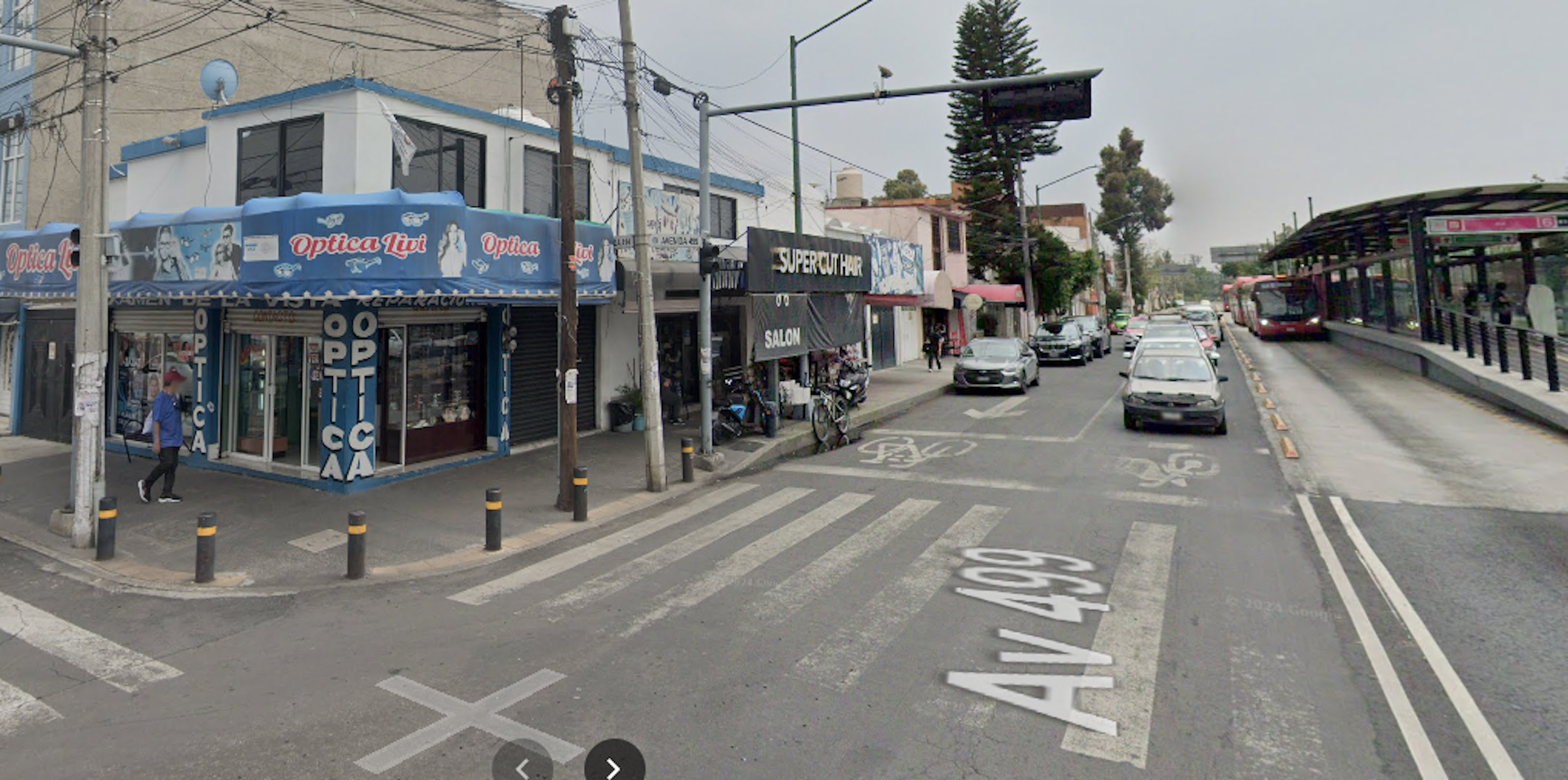 La imagen muestra una calle comercial urbana con diversos negocios, incluyendo una óptica en la esquina con fachada azul y blanca. Se observa una mezcla de edificios comerciales de baja altura, vehículos estacionados, y una parada de autobús. La calle tiene señalizaciones viales, un cruce peatonal y carriles para bicicletas, indicando una infraestructura urbana moderna.