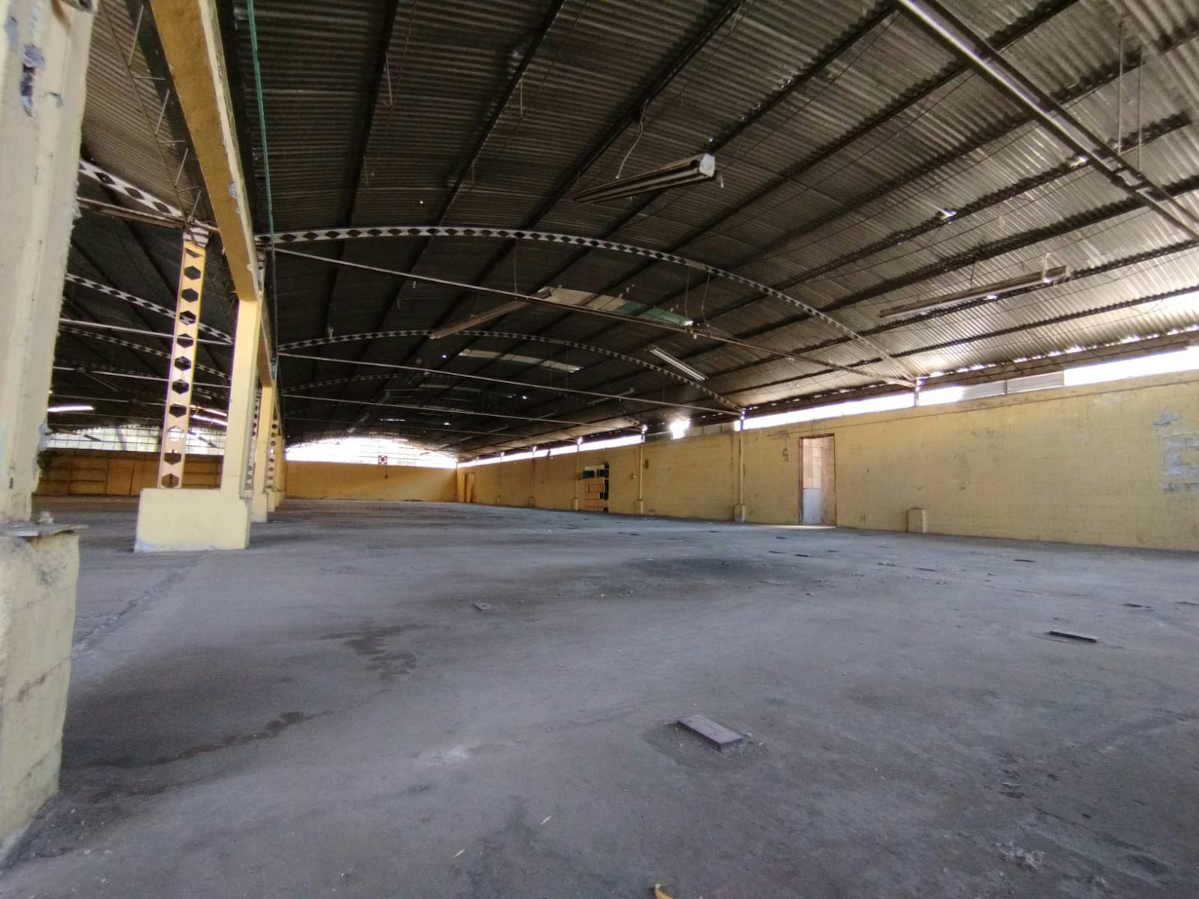 Esta imagen muestra un gran espacio industrial vacío. Tiene un techo de metal corrugado con vigas expuestas y paredes amarillas. El piso es de concreto y hay una puerta visible al fondo. El espacio parece estar en desuso o abandonado, con algunas manchas y suciedad visibles. Este tipo de espacio podría ser ideal para una reconversión en lofts, almacenes o espacios comerciales amplios.