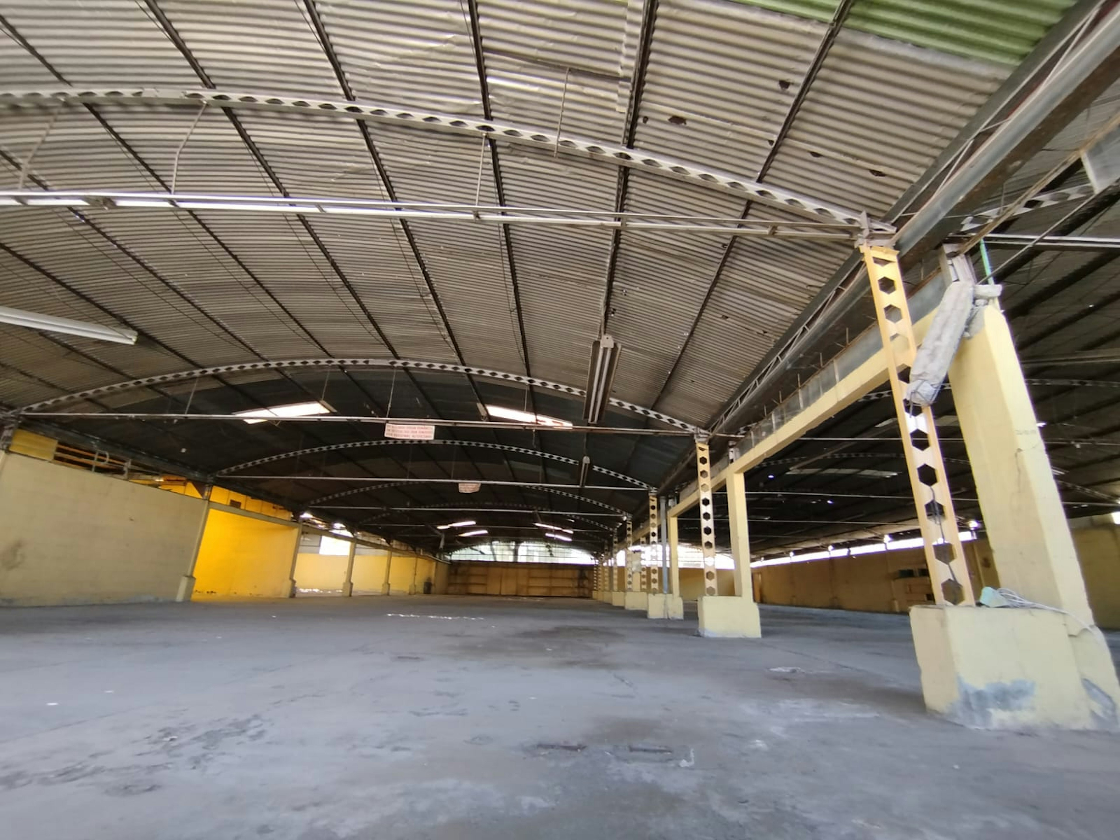 Esta imagen muestra un amplio espacio industrial o de almacenamiento vacío. El techo es alto y arqueado, con vigas metálicas visibles y paneles corrugados. Las paredes son en parte amarillas y blancas. El suelo es de concreto y está despejado. Se pueden ver columnas y estructuras de soporte. El espacio parece estar en desuso o en proceso de renovación, ideal para ser adaptado a diversos usos comerciales o industriales.