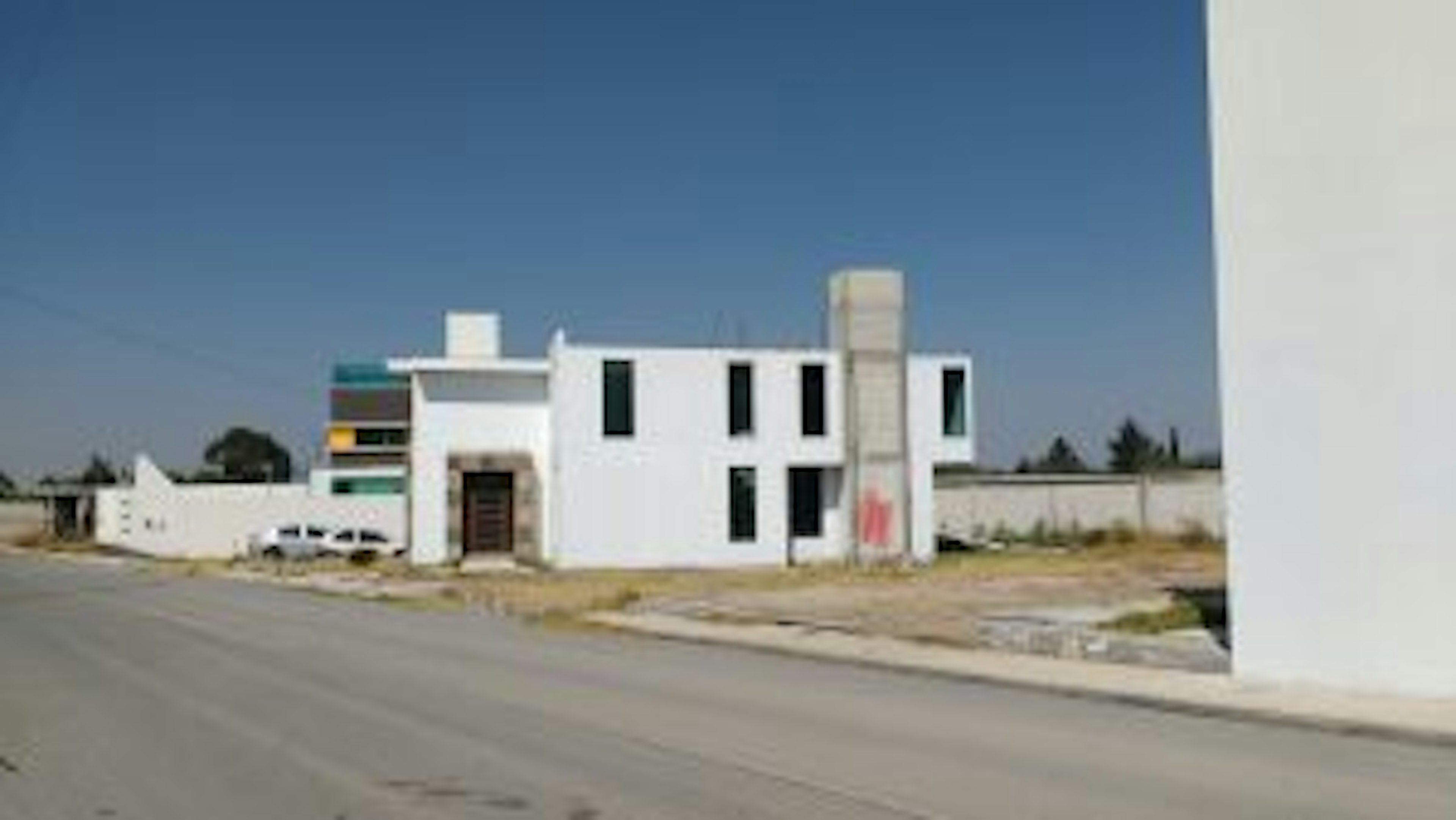 La imagen muestra una casa moderna de dos pisos con un diseño minimalista. La fachada es principalmente blanca con ventanas negras rectangulares. Tiene un acceso principal con una puerta oscura y un elemento vertical alto, posiblemente decorativo o funcional. La propiedad está situada en un terreno amplio con una calle pavimentada al frente. El cielo es azul claro, sugiriendo un clima soleado. La arquitectura es contemporánea y la casa parece estar en una zona residencial en desarrollo.