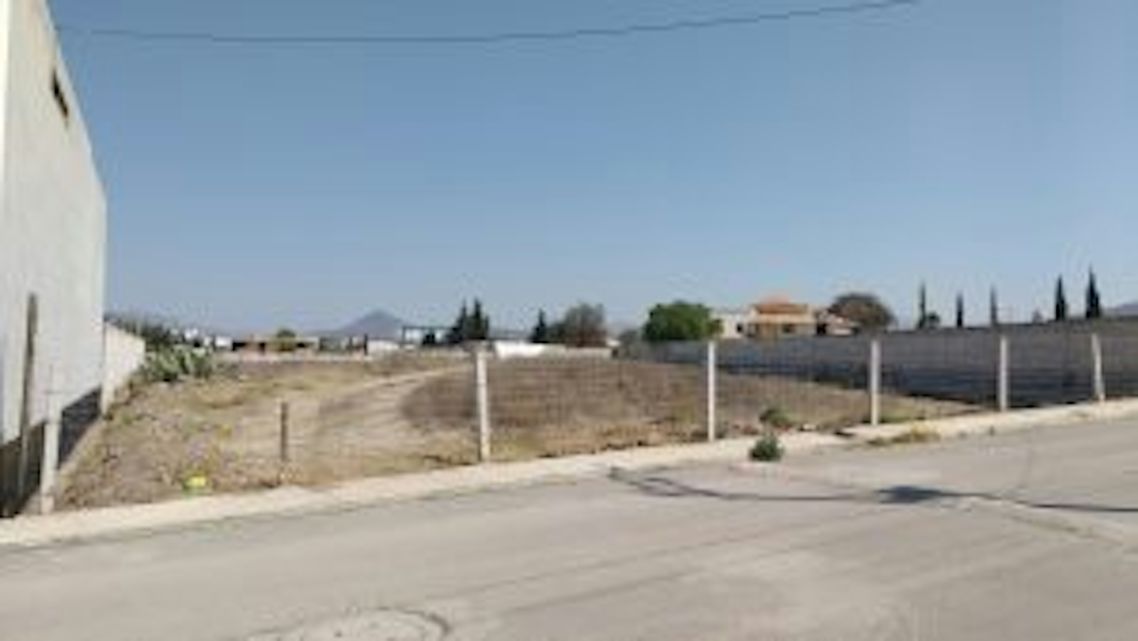 La imagen muestra un terreno baldío en una zona suburbana o rural. Se observa un área sin desarrollar con vegetación escasa, delimitada por postes y muros. Al fondo se ven algunas construcciones y árboles. El cielo es claro y despejado. El terreno parece estar en una zona de desarrollo potencial, ideal para construcción residencial o comercial.