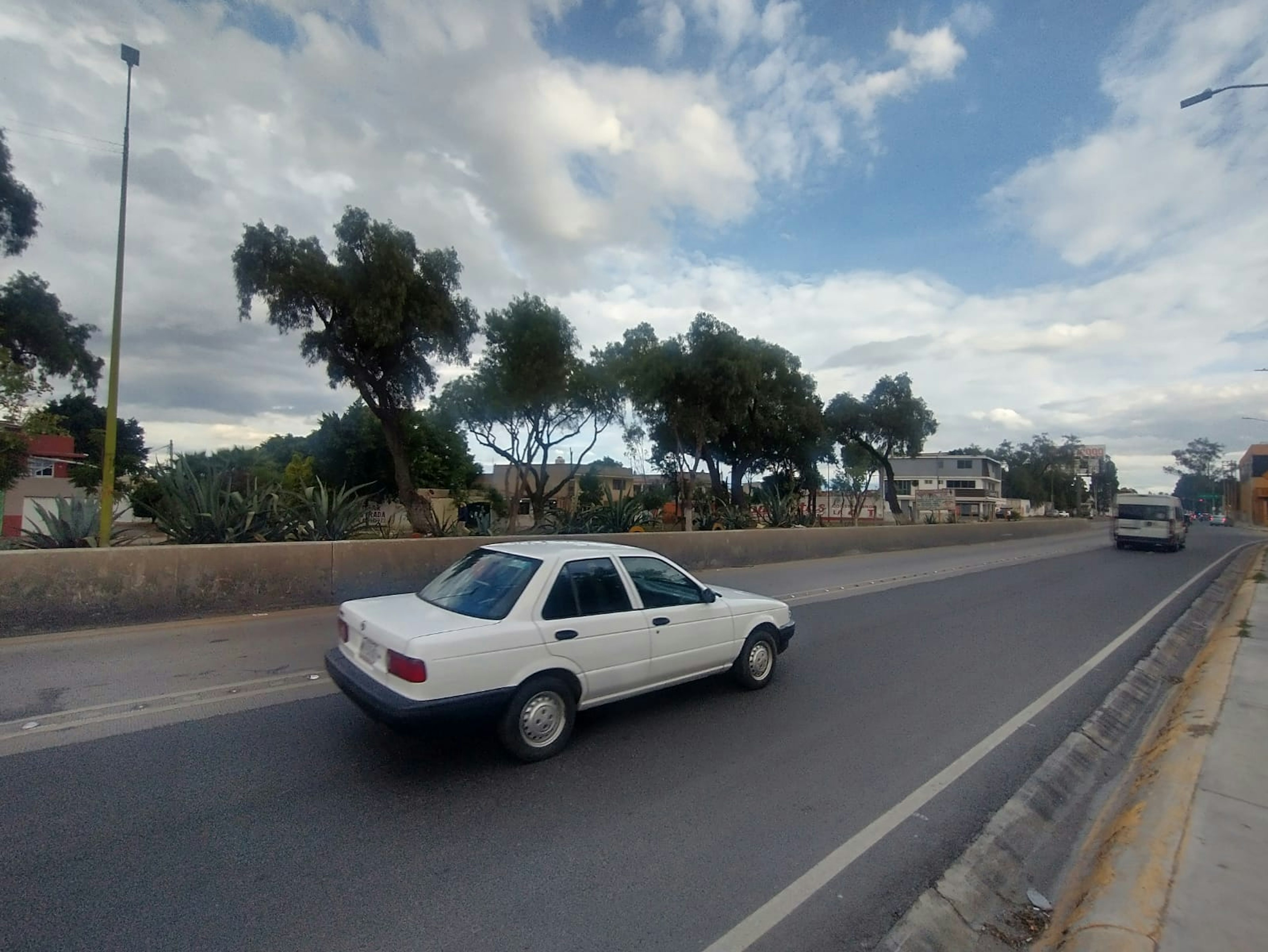 La imagen muestra una calle amplia con un vehículo blanco estacionado al lado. Al fondo, se observan casas y vegetación, indicando una zona residencial posiblemente en las afueras de una ciudad.