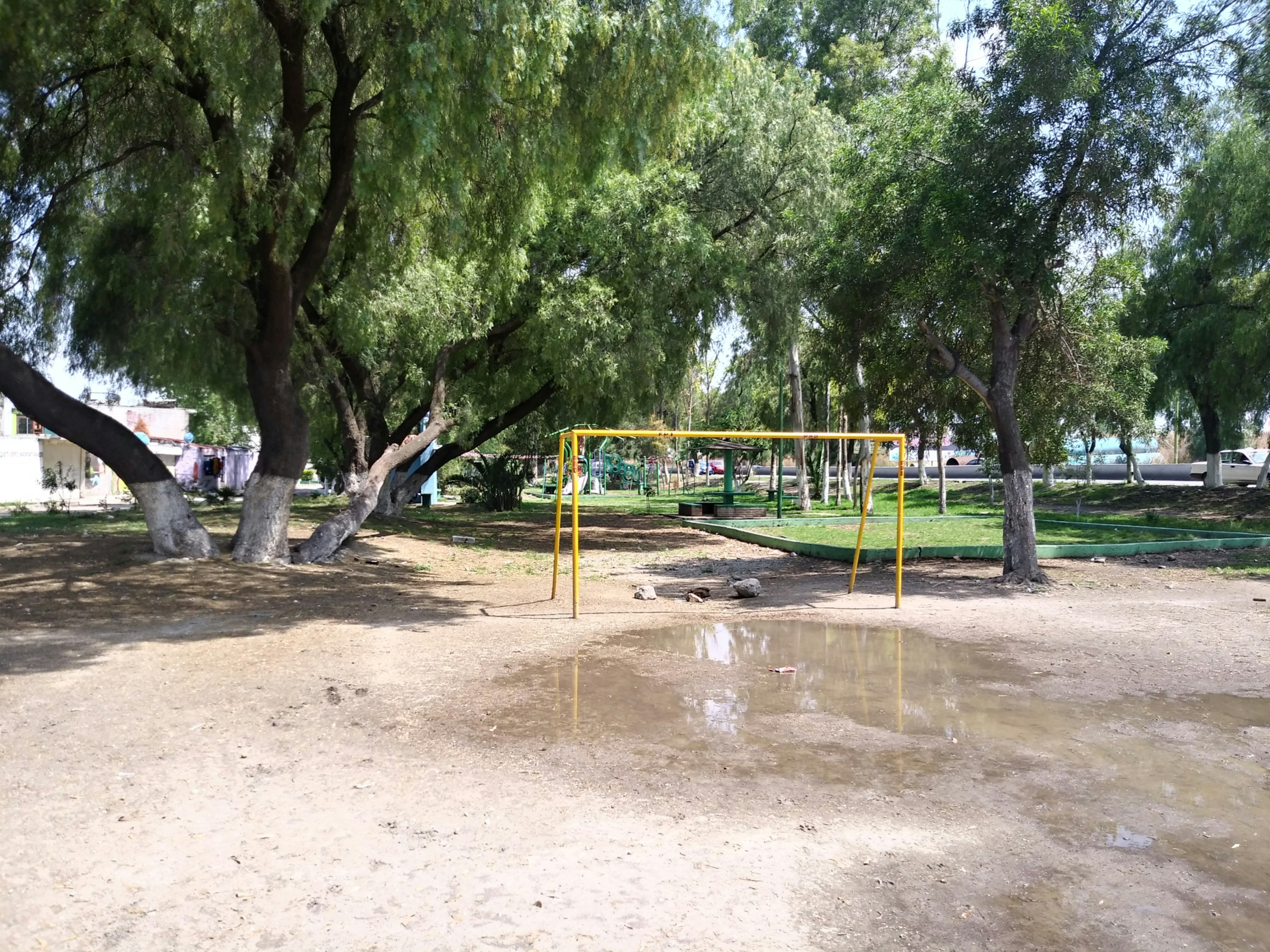 La imagen muestra un parque público con áreas verdes y juegos infantiles. Los árboles grandes proporcionan sombra y un ambiente natural, ideal para familias y actividades al aire libre.