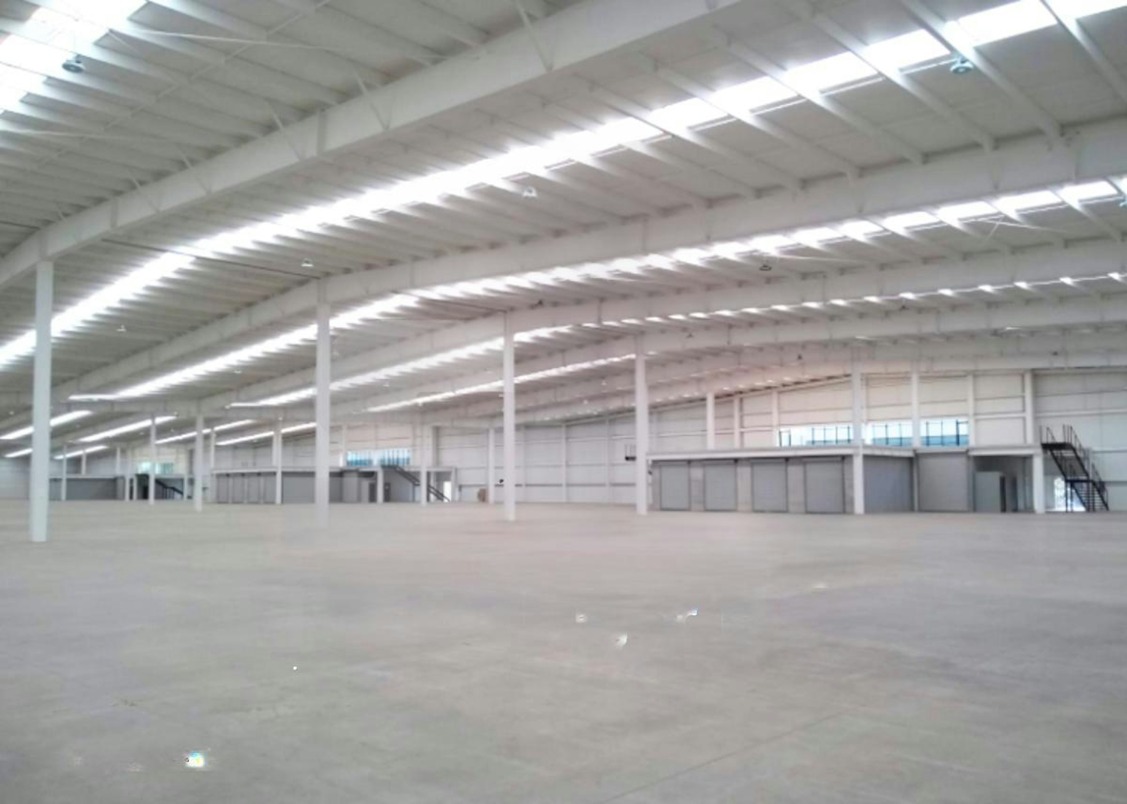 La imagen muestra un amplio espacio industrial interior, posiblemente un almacén o centro de distribución. Tiene un techo alto con vigas expuestas y una iluminación brillante. El piso es de concreto liso y hay algunas estructuras elevadas visibles. El espacio está vacío, dando una sensación de amplitud. En la parte inferior de la imagen se puede ver parte de un logo o texto que dice INDUSTRY, sugiriendo que es una instalación industrial o logística.