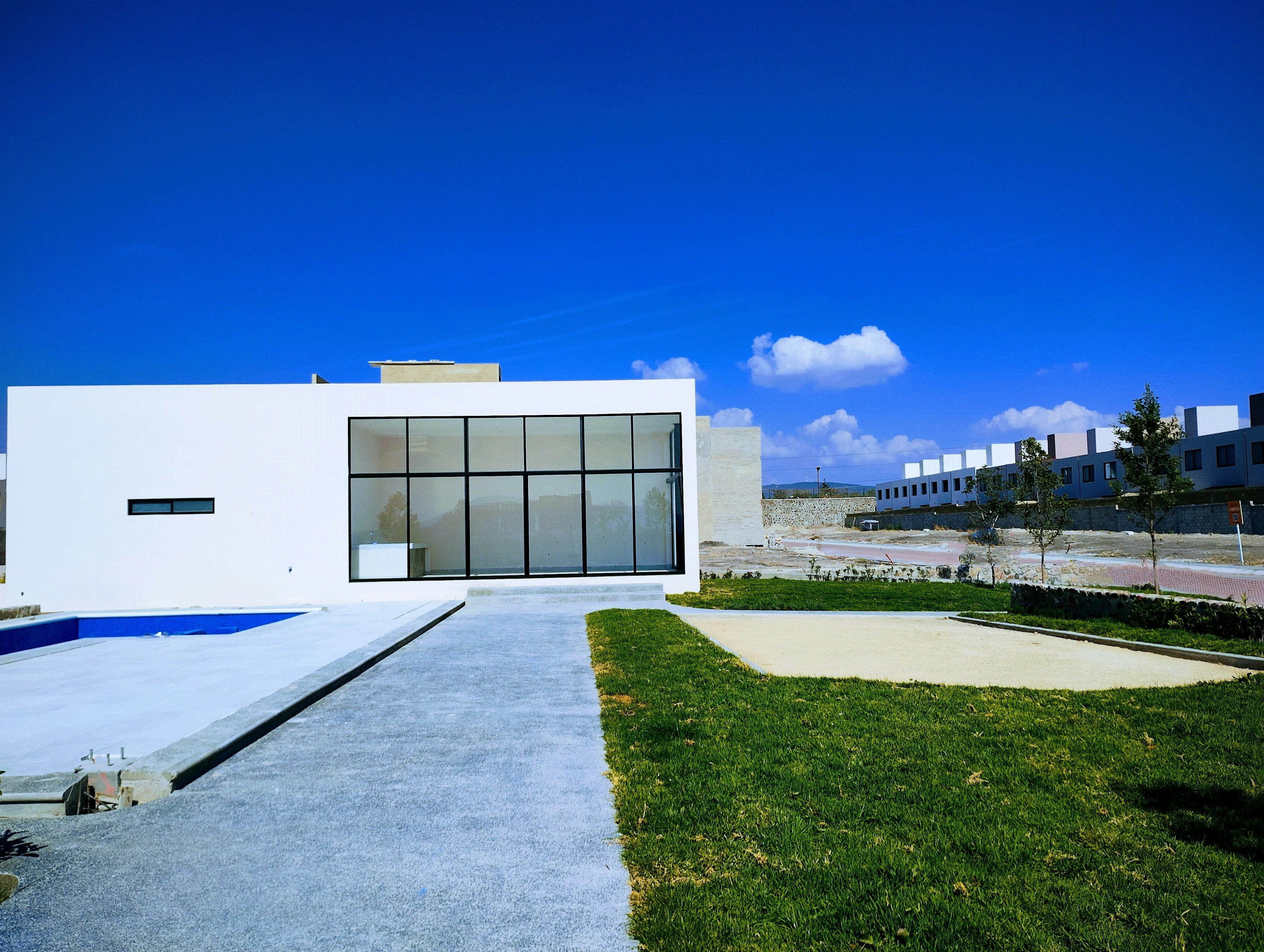 La imagen muestra una moderna casa de diseño minimalista con grandes ventanales y líneas limpias. La fachada es blanca y cuenta con una piscina en el frente. El paisajismo incluye césped bien cuidado y un camino de concreto. Al fondo se observan otras propiedades, sugiriendo que es parte de un desarrollo residencial exclusivo. El cielo azul brillante y la arquitectura contemporánea crean una atmósfera de lujo y sofisticación.