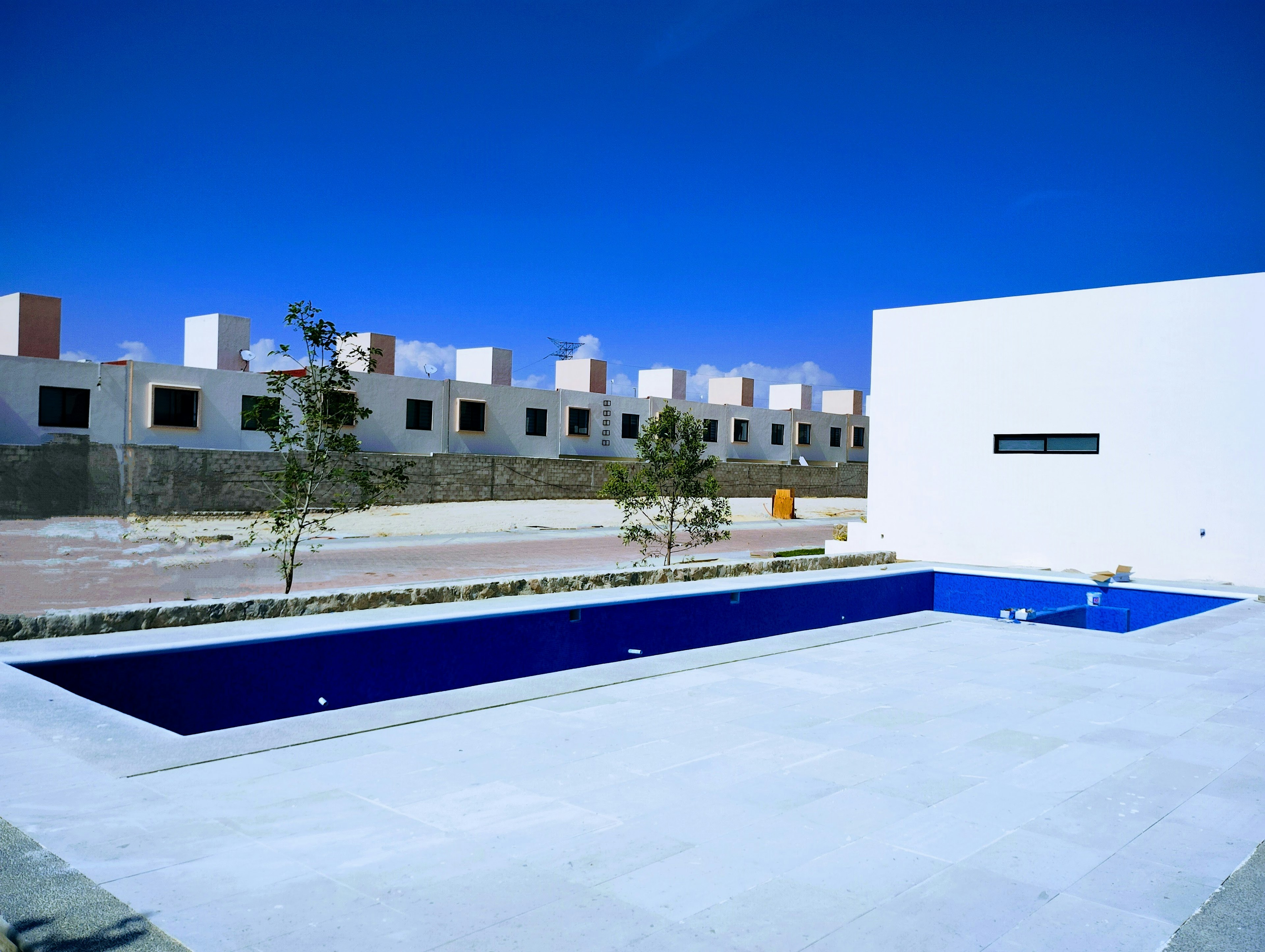 La imagen muestra un complejo residencial moderno con edificios blancos de estilo minimalista. En primer plano se aprecia una piscina rectangular de color azul intenso rodeada de un área de concreto. Al fondo se observa una hilera de casas o apartamentos con pequeñas ventanas y techos planos. El cielo es de un azul brillante, sugiriendo un clima cálido y soleado. El diseño general es limpio y geométrico, típico de desarrollos inmobiliarios contemporáneos.