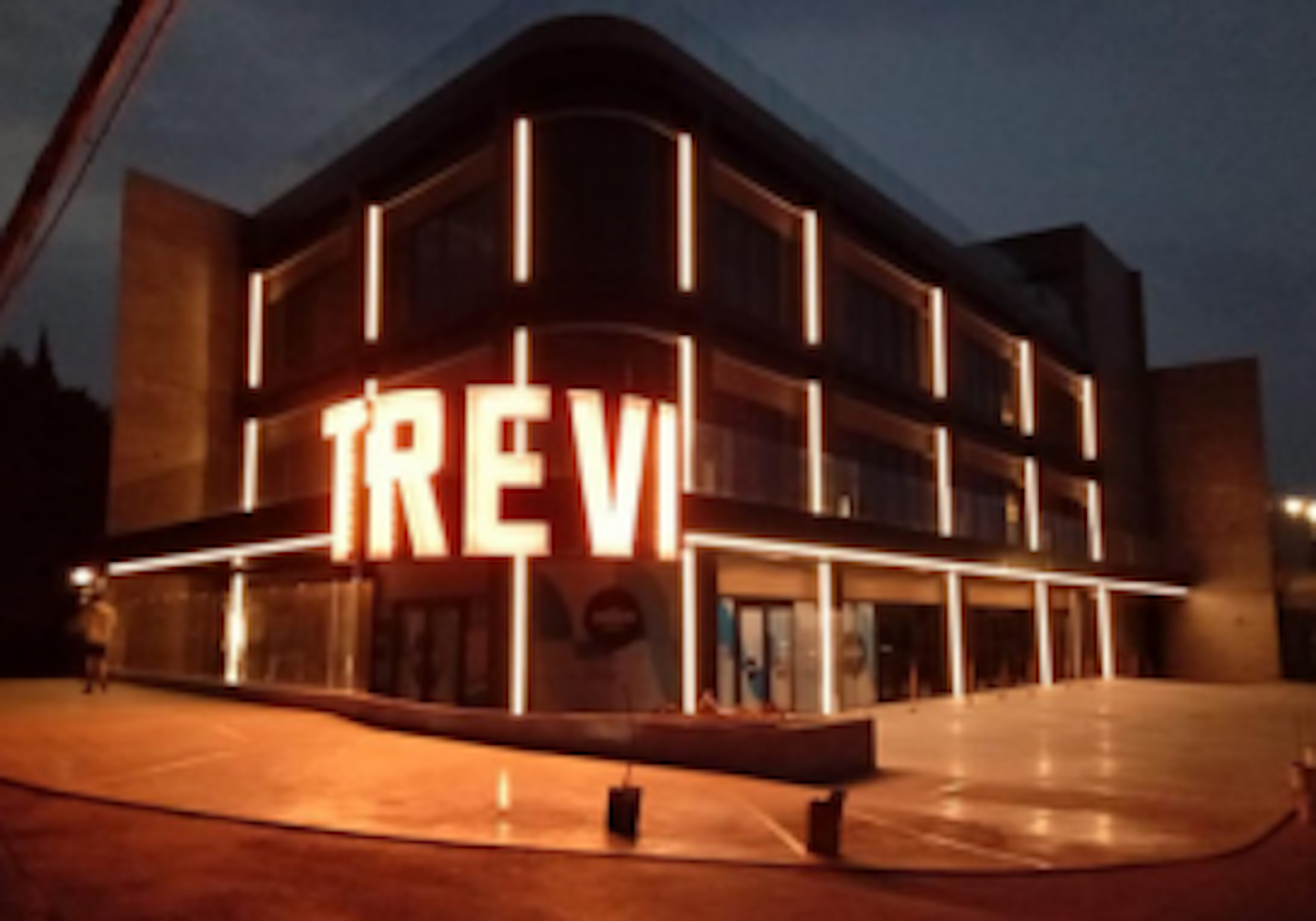 Una impresionante fachada moderna de un edificio iluminado por la noche, con el nombre TREV en grandes letras luminosas. La estructura combina elementos arquitectónicos contemporáneos con una iluminación estratégica que resalta sus líneas y formas. El diseño sugiere un espacio comercial o de entretenimiento de alta gama, posiblemente un teatro, cine o centro cultural vanguardista.