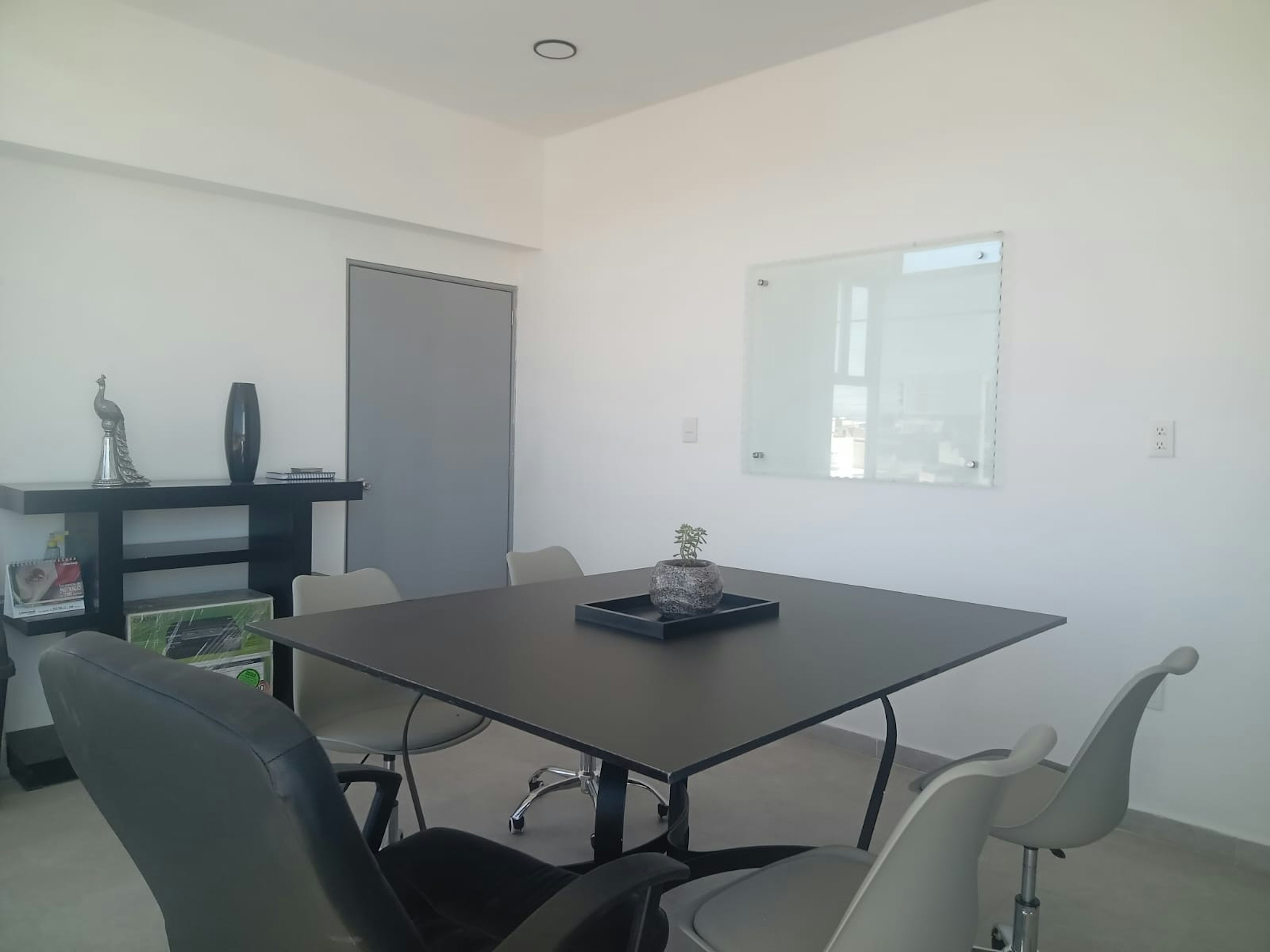 Esta imagen muestra una sala de reuniones o espacio de oficina moderno y minimalista. Tiene una mesa cuadrada negra en el centro con sillas blancas y una silla de oficina negra. Hay una pizarra blanca en la pared, un estante decorativo con un jarrón y una figura de pavo real. La habitación tiene paredes blancas, un techo con iluminación empotrada y una puerta gris. El diseño es limpio y funcional, ideal para un entorno profesional.