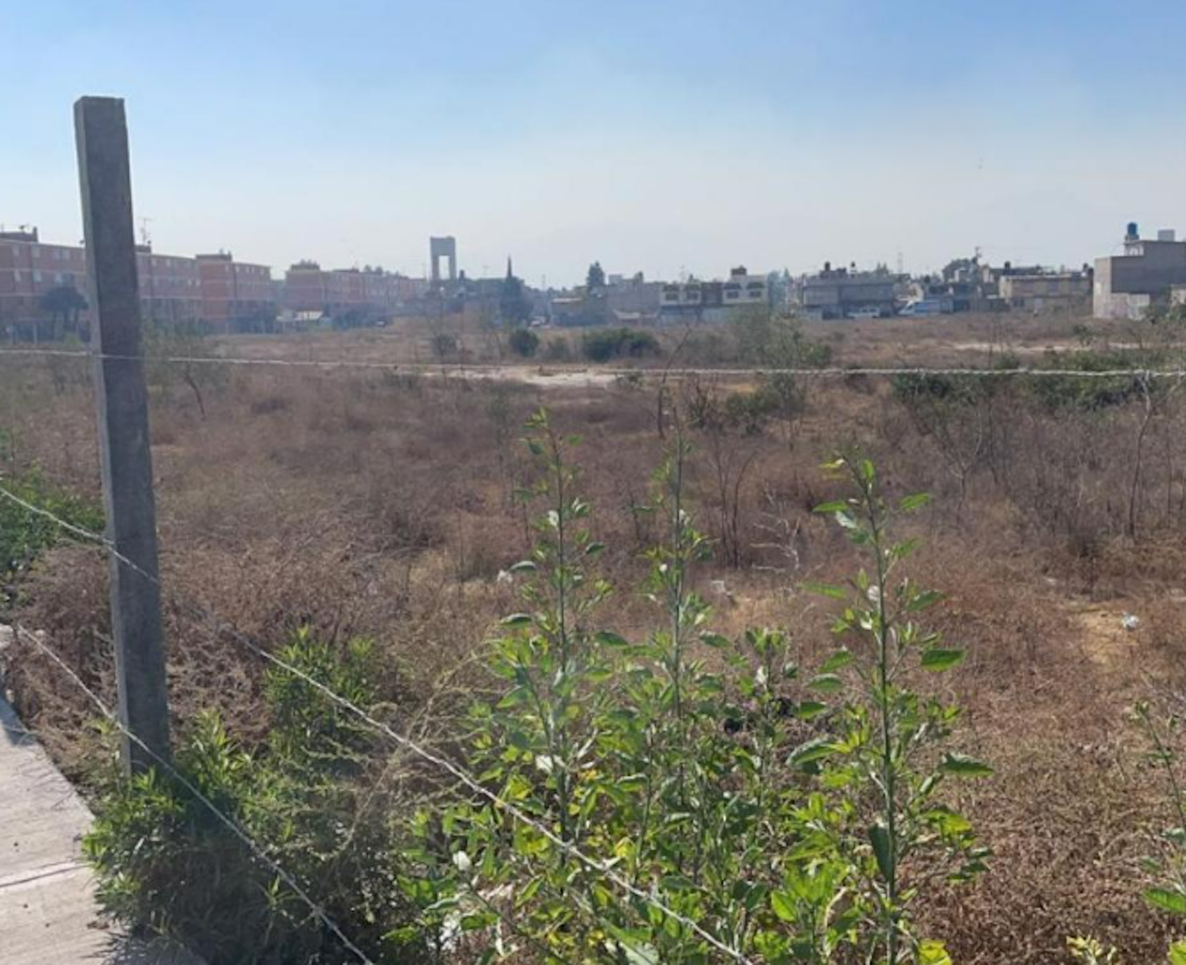 La imagen muestra un terreno baldío en una zona urbana. En primer plano se ve un área descuidada con vegetación seca y algunos arbustos verdes. Al fondo se aprecian edificios residenciales y comerciales, indicando que es un área en desarrollo. El terreno parece estar cercado, sugiriendo que podría ser una propiedad privada lista para futuros proyectos inmobiliarios.