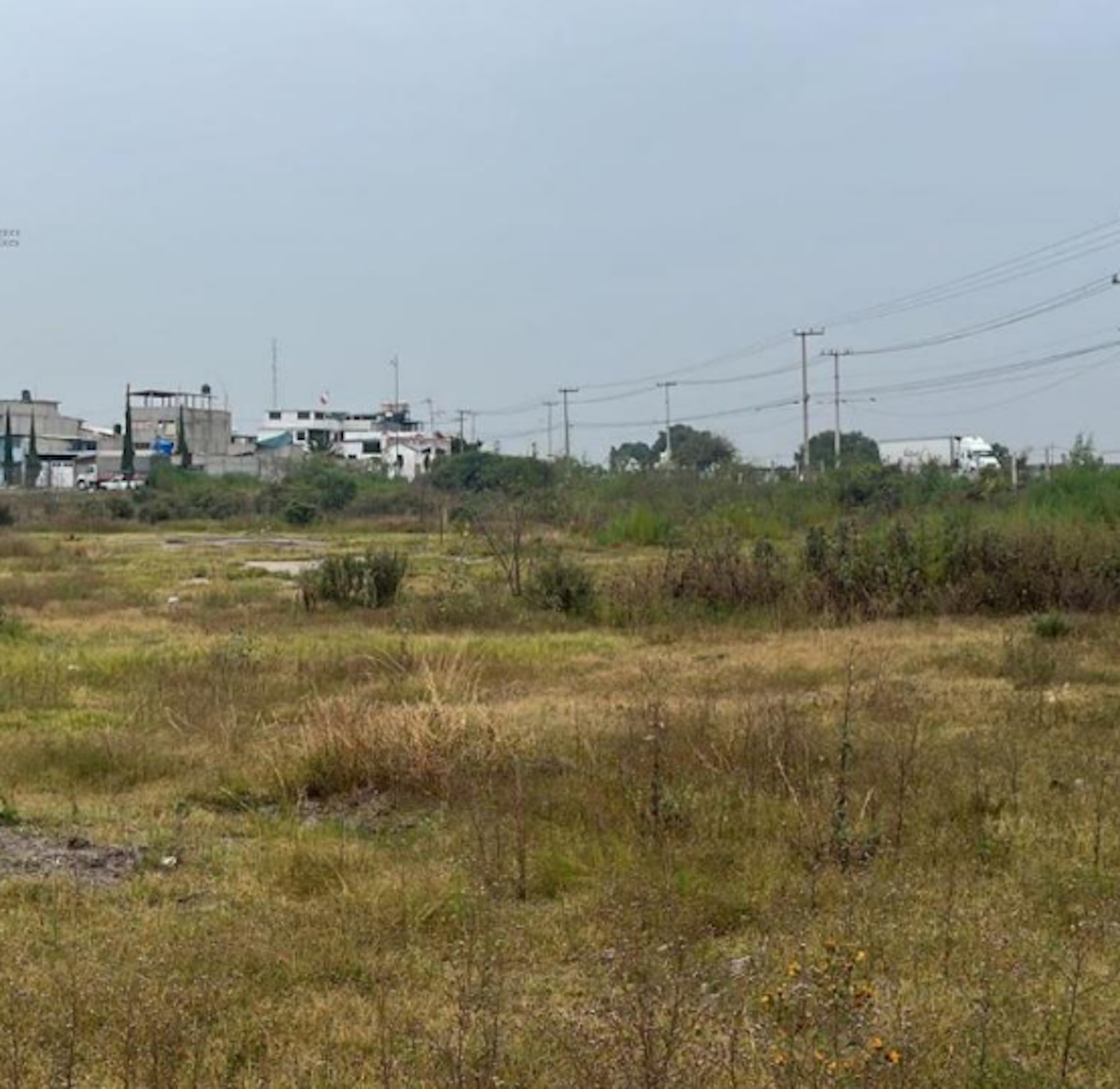 La imagen muestra un terreno baldío en primer plano, con vegetación silvestre y seca. Al fondo se observa una zona urbana con construcciones sencillas, postes de electricidad y cables. El área parece ser periférica, con potencial para desarrollo inmobiliario debido a la presencia de infraestructura básica y espacio disponible para nuevas edificaciones.