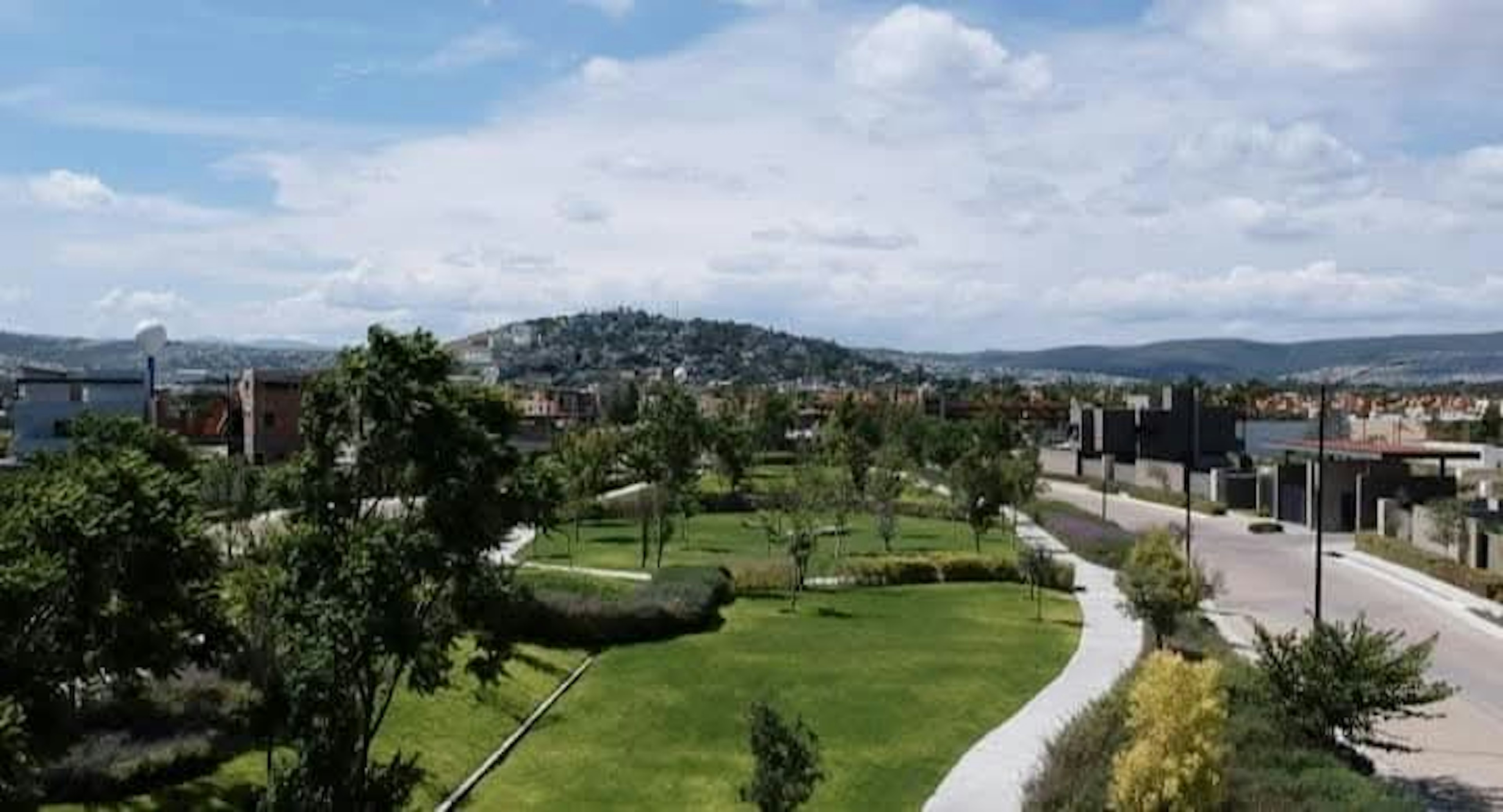 La imagen muestra una vista panorámica de una zona residencial con un parque en primer plano. Se observa un área verde bien cuidada con caminos pavimentados, árboles y arbustos. Al fondo se aprecian casas y edificios, así como colinas boscosas. El cielo es azul con algunas nubes. La escena sugiere un desarrollo inmobiliario planificado con espacios verdes integrados, ofreciendo un entorno agradable y tranquilo para vivir.