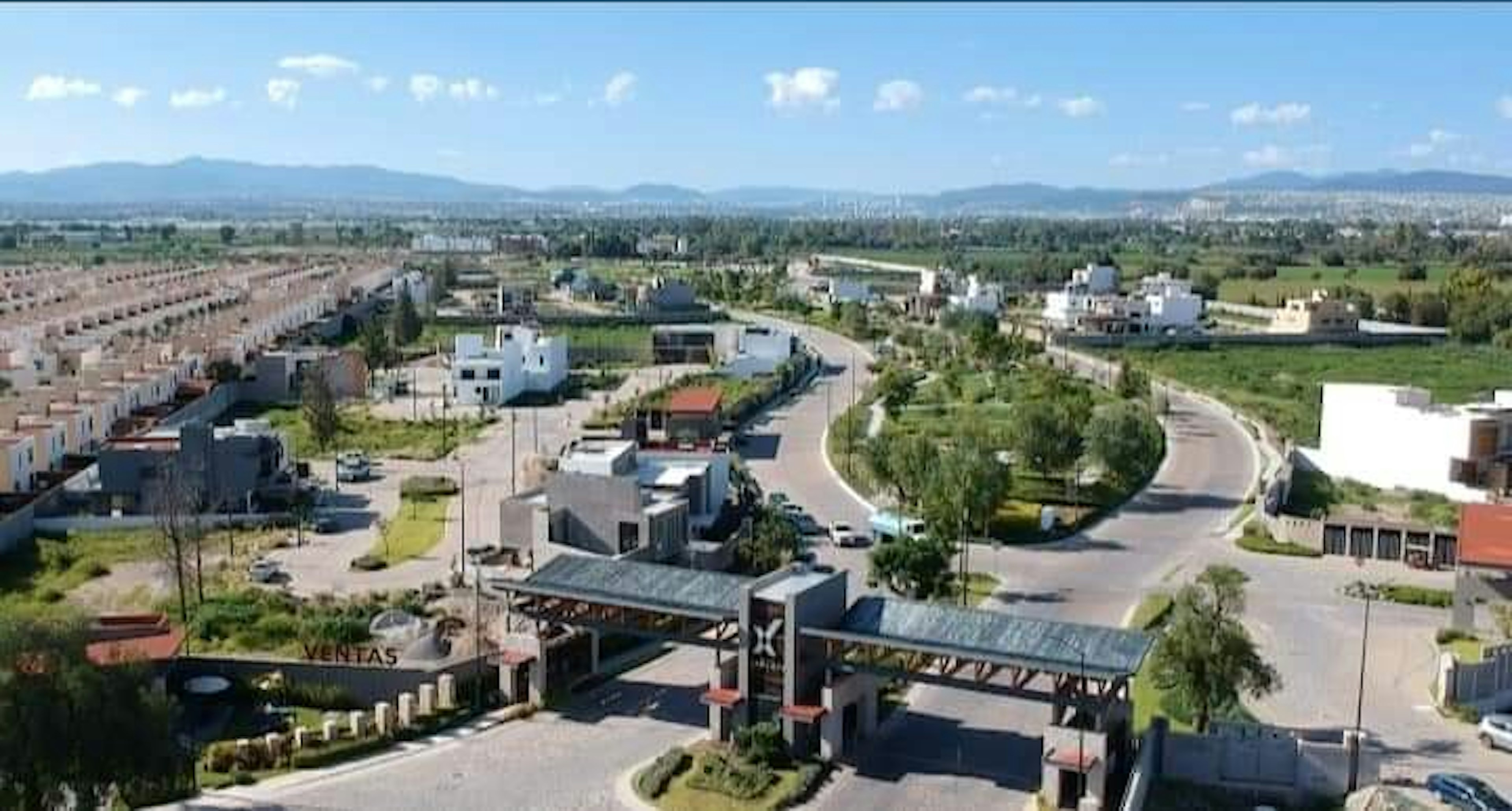 La imagen muestra una vista aérea de un desarrollo inmobiliario en expansión. Se observa una mezcla de viviendas unifamiliares y edificios más grandes, con calles curvas y áreas verdes. Hay una entrada principal con una estructura cubierta, posiblemente un área de ventas. El paisaje circundante incluye montañas en el horizonte y vegetación dispersa. El desarrollo parece estar en una etapa intermedia de construcción, con algunas áreas completadas y otras en progreso.