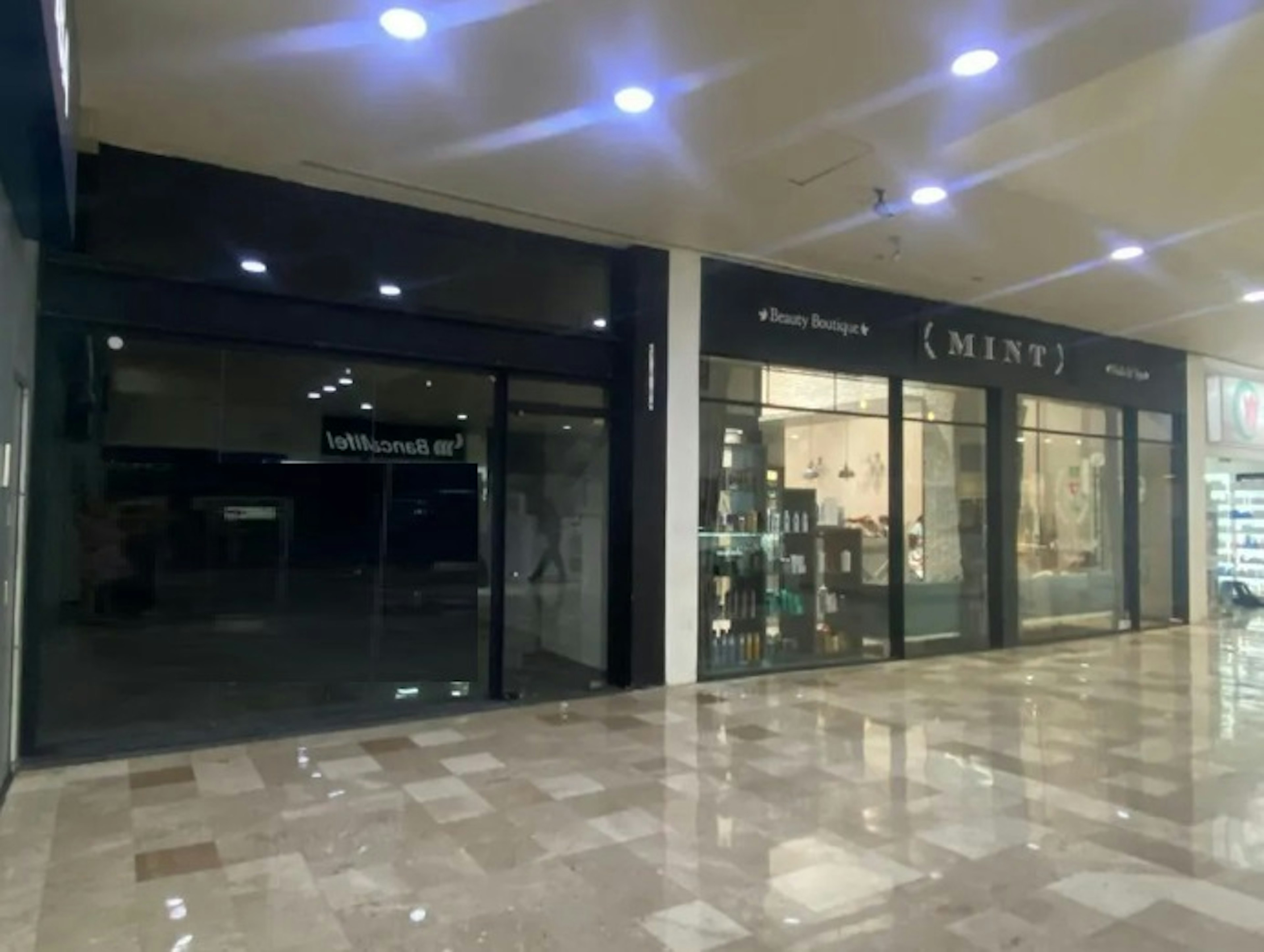 La imagen muestra el interior de un centro comercial o galería comercial. Se pueden ver dos locales comerciales: uno con un letrero negro que parece estar cerrado o vacío, y otro llamado MINT que parece ser una tienda de belleza o boutique. El suelo es de mármol brillante y reflectante, y el techo tiene luces empotradas que crean una atmósfera moderna y luminosa. El espacio parece estar bien mantenido y listo para albergar negocios.