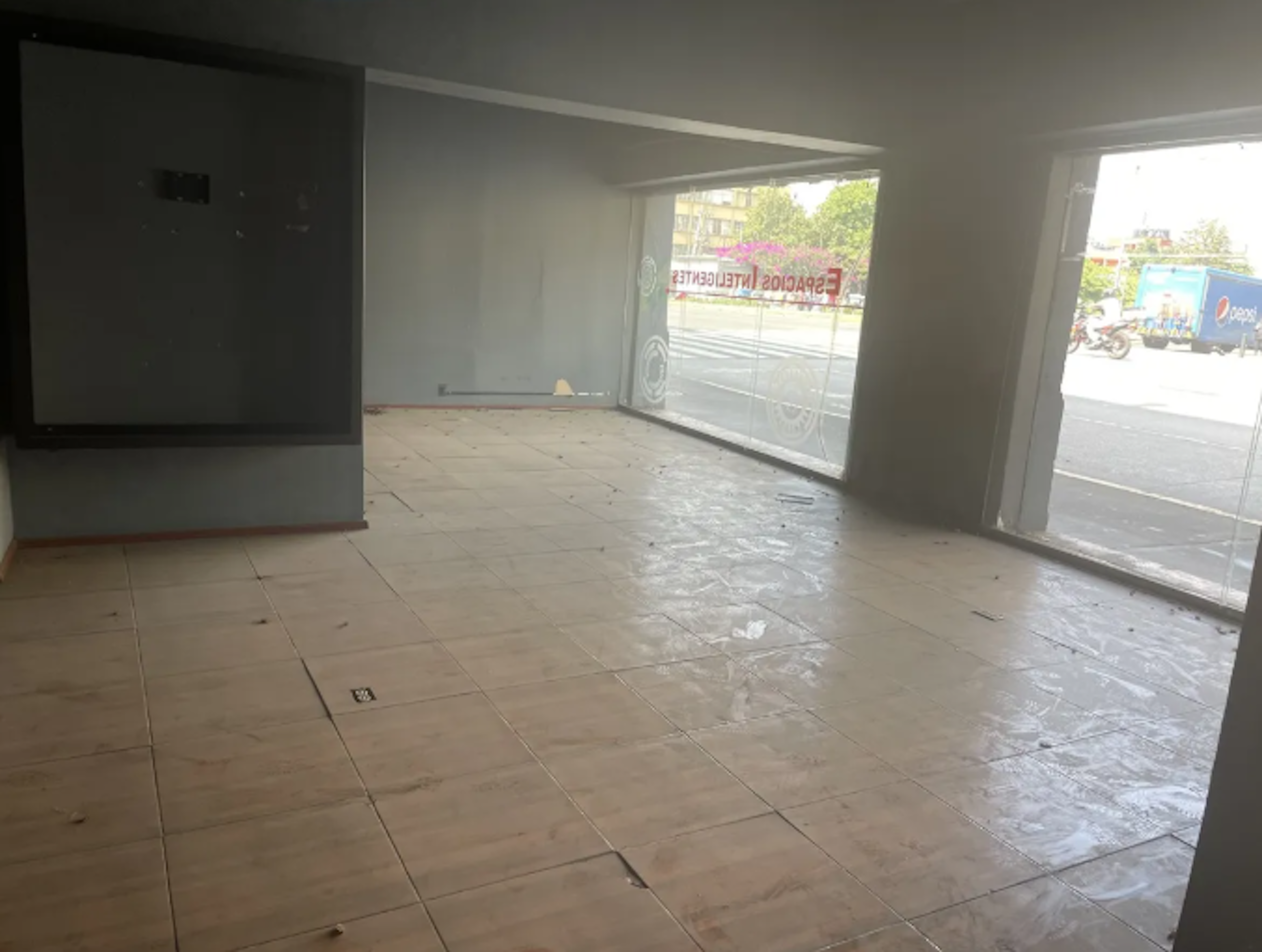 La imagen muestra un local comercial vacío en proceso de renovación. Tiene un amplio espacio interior con piso de baldosas y grandes ventanales que dan a la calle. Se puede ver un letrero exterior reflejado en el vidrio. El local parece estar en una etapa temprana de remodelación, con paredes blancas y algunas áreas del piso aún sin terminar. Es un espacio luminoso con potencial para diversos usos comerciales.