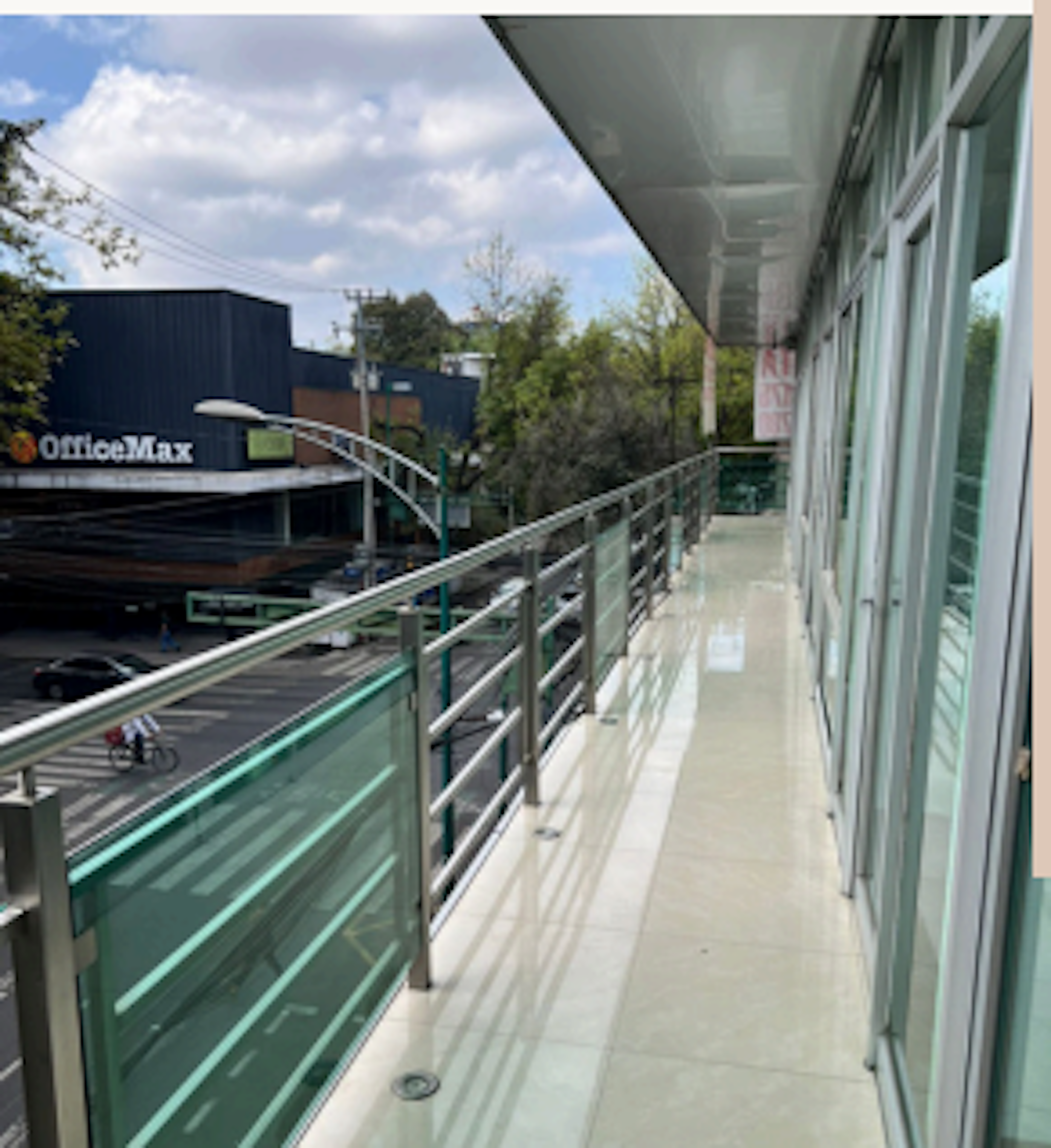 La imagen muestra una vista desde un balcón o terraza de un edificio comercial moderno. Se observa una barandilla de metal y vidrio verde, y una amplia acera. Al otro lado de la calle se ve un edificio de OfficeMax. El entorno incluye árboles y un cielo parcialmente nublado, sugiriendo una zona comercial urbana con espacios de oficinas y tiendas.