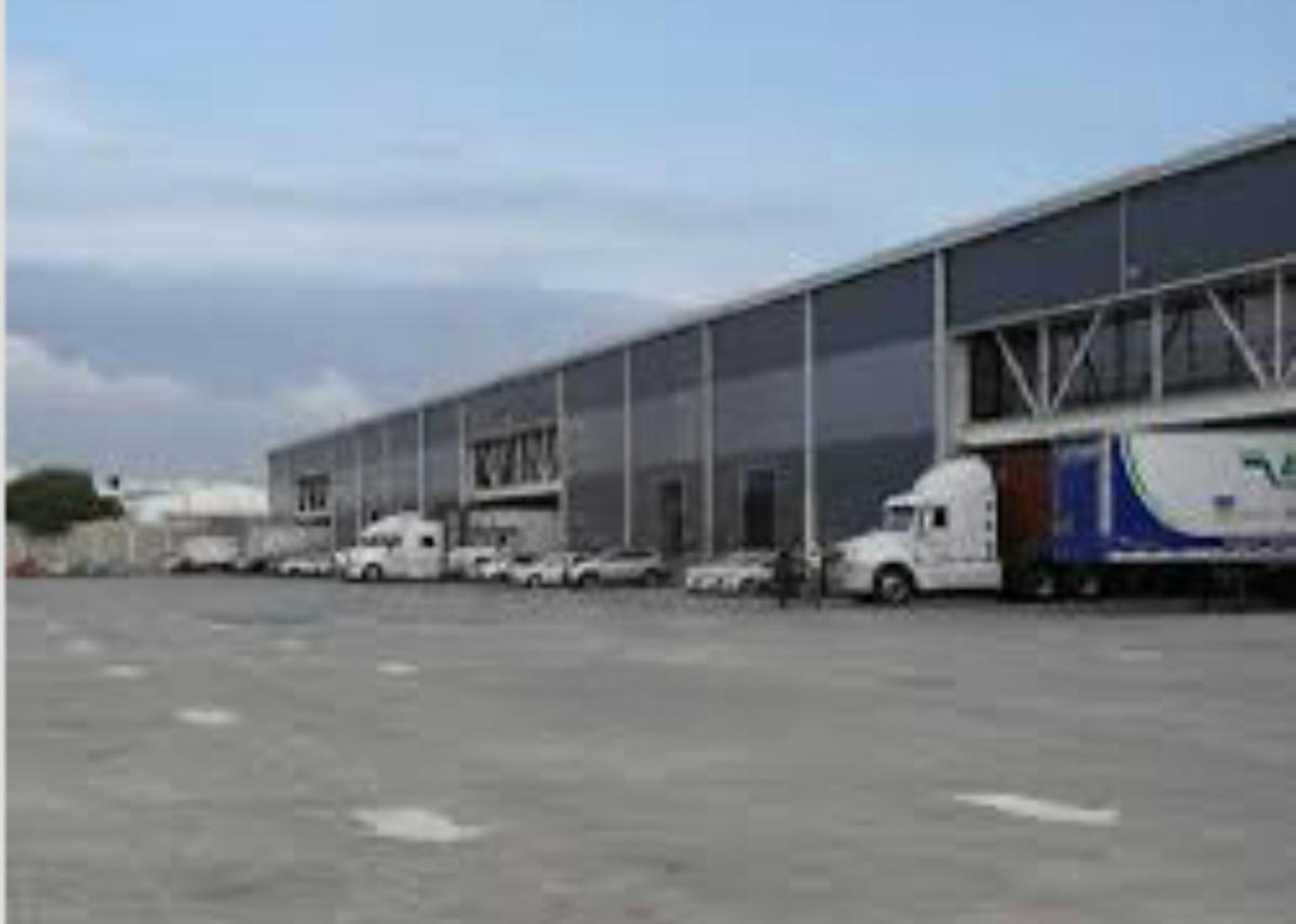 La imagen muestra un complejo industrial con grandes almacenes o naves y camiones estacionados. Se ve una estructura moderna con techos altos y amplias entradas para vehículos de carga. El primer plano muestra un área pavimentada, sugiriendo un patio de maniobras. Hay un logotipo superpuesto que dice Industry y menciona Strategic, Industry y Logistics, indicando que es una instalación logística o de distribución industrial.