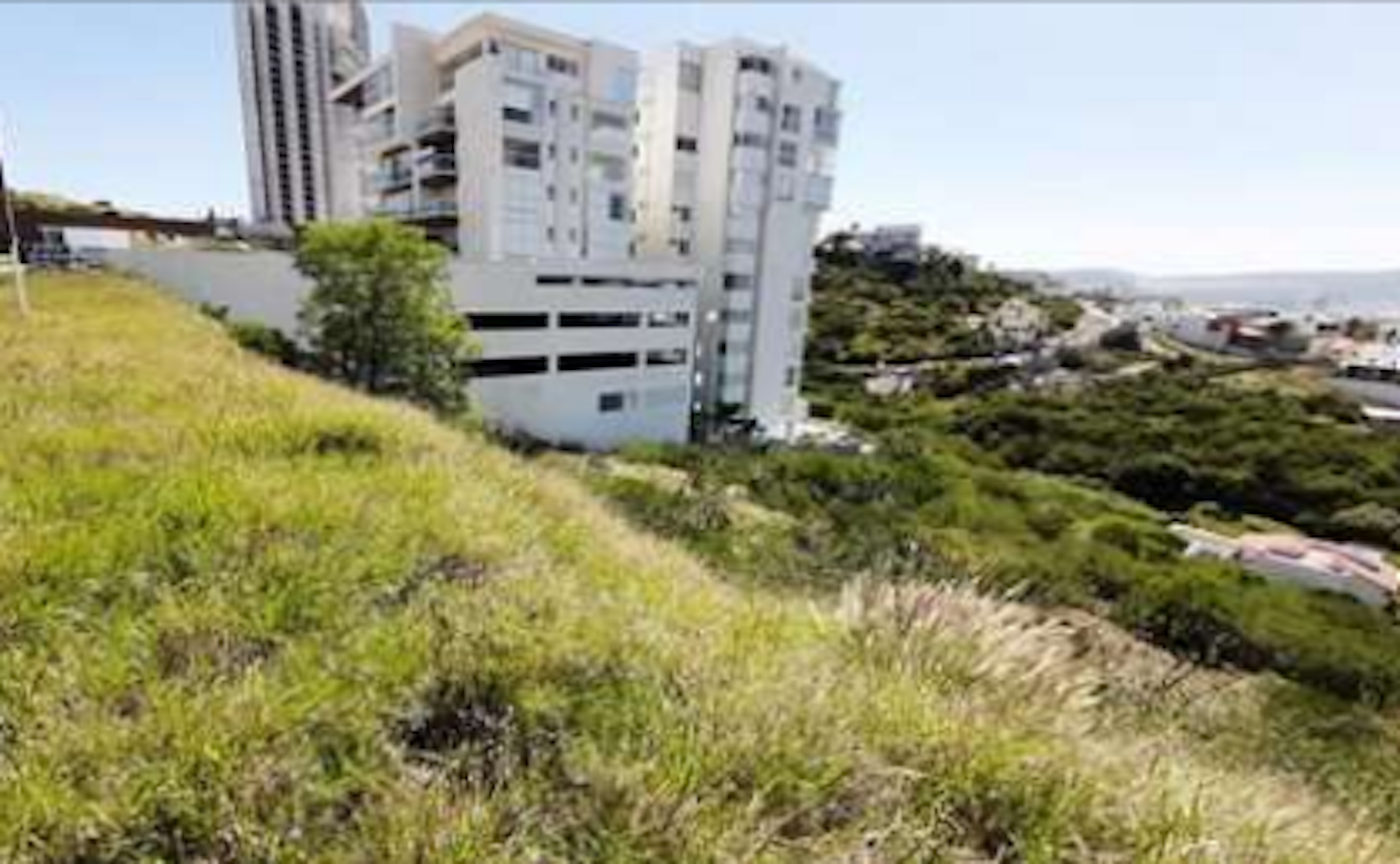 La imagen muestra un paisaje urbano con edificios residenciales modernos en una colina. En primer plano se observa una zona verde con césped y vegetación. Los edificios son de color claro, con varios pisos y balcones. Se aprecia un estacionamiento de varios niveles junto a los edificios. Al fondo se vislumbra más vegetación y lo que parece ser una vista panorámica de la ciudad. El entorno sugiere un desarrollo inmobiliario en una zona elevada con vistas.