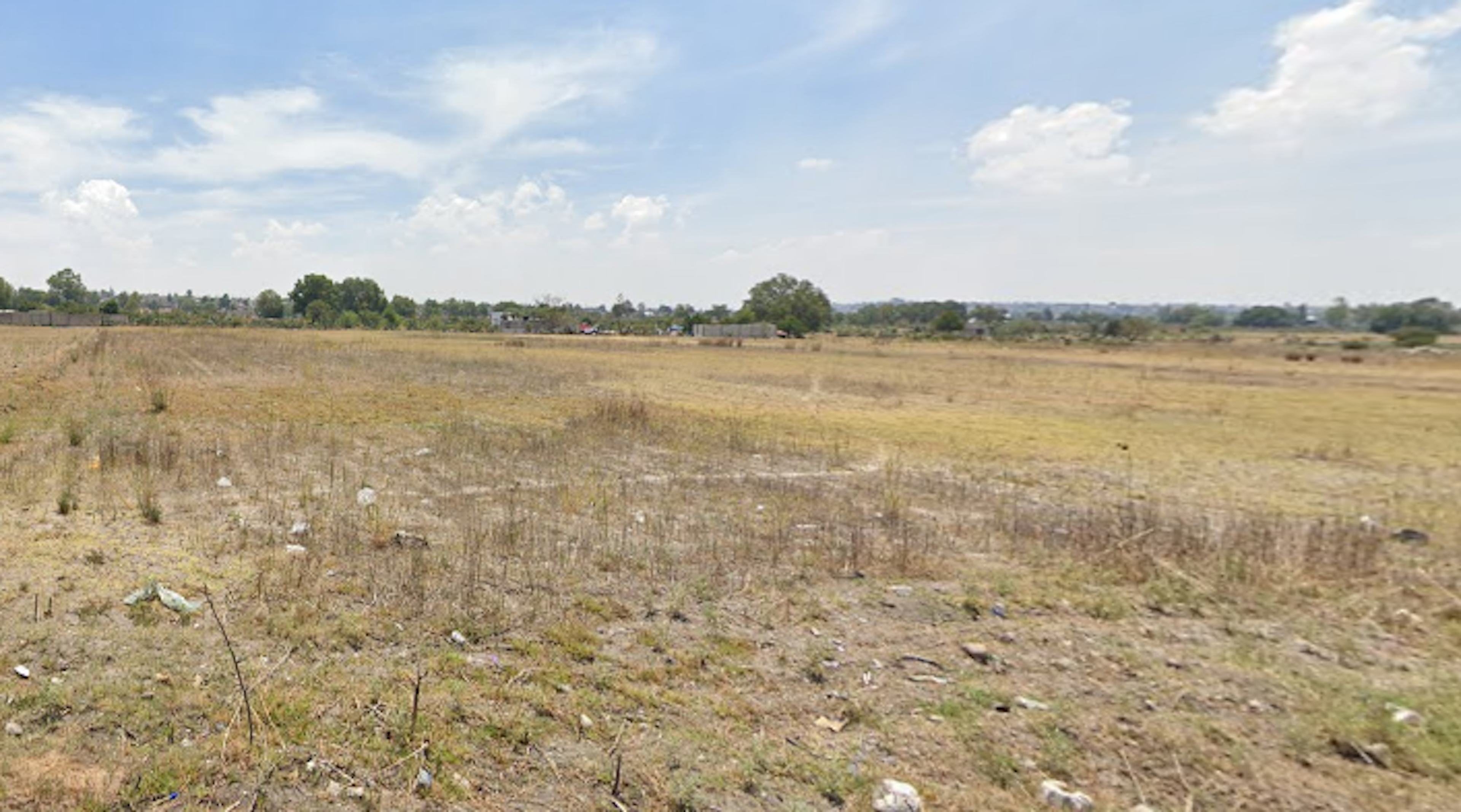 La imagen muestra un terreno amplio y plano, aparentemente sin desarrollar, con vegetación escasa y seca. El paisaje es rural, con árboles y algunas estructuras visibles en el horizonte. El cielo es claro con algunas nubes. El terreno parece ideal para desarrollo inmobiliario, ofreciendo un espacio extenso con potencial para construcción residencial o comercial en un entorno suburbano o rural.