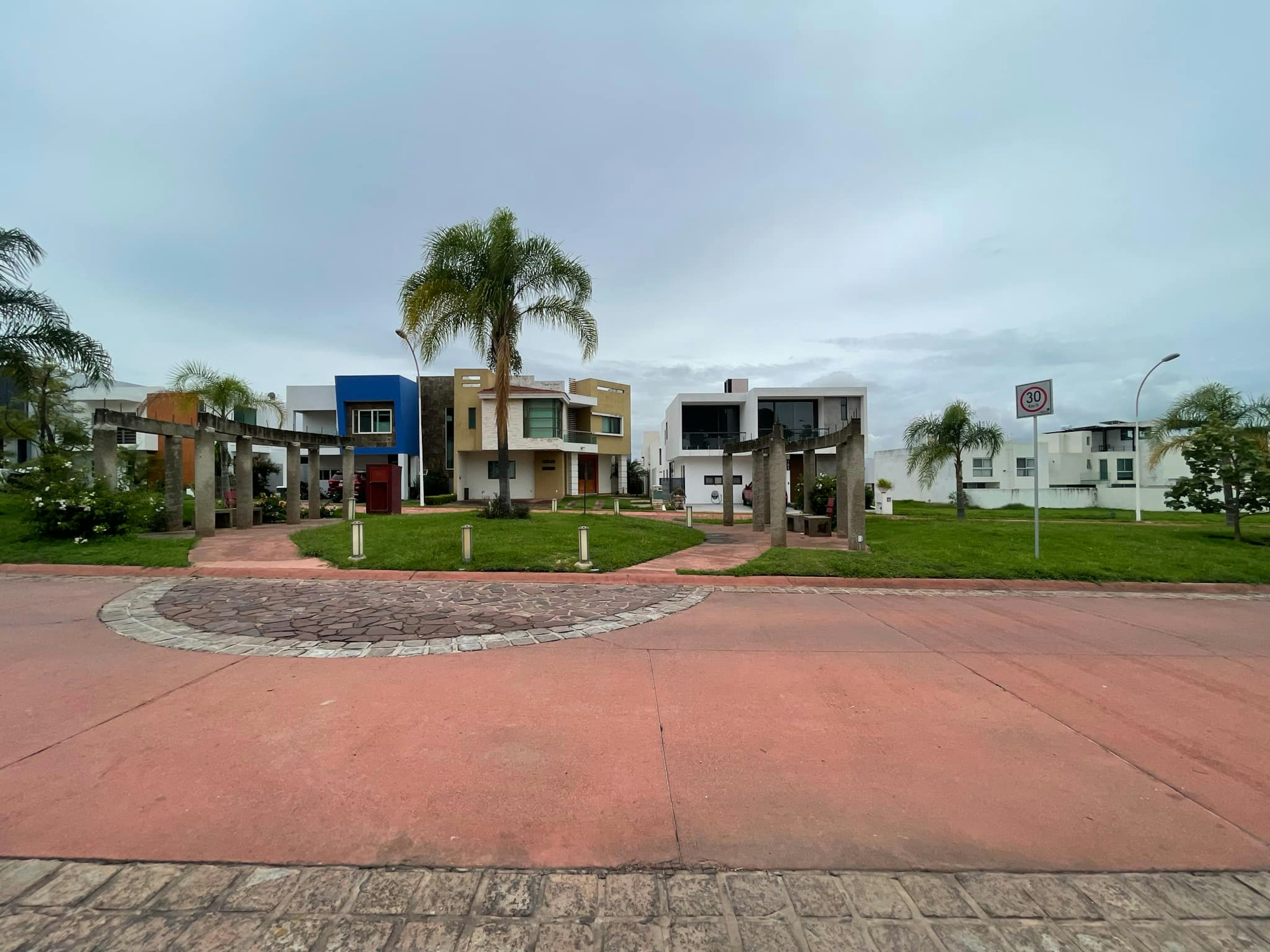 La imagen muestra un moderno complejo residencial con viviendas de diseño contemporáneo. Las casas tienen fachadas coloridas y estilizadas, con detalles arquitectónicos únicos. El área común incluye un sendero pavimentado, áreas verdes bien cuidadas y palmeras. El ambiente sugiere una comunidad planificada de alto nivel, ideal para familias o profesionales que buscan un estilo de vida cómodo y elegante en un entorno tranquilo y seguro.