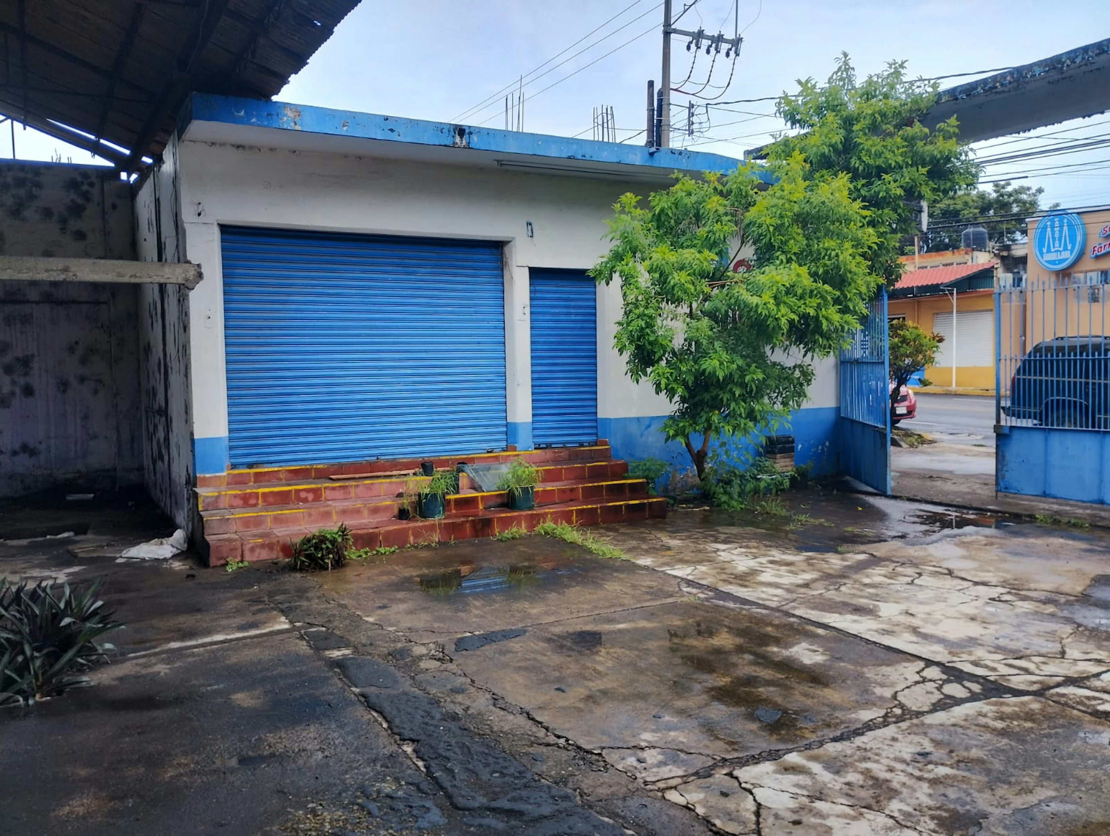 La imagen muestra una propiedad comercial en una zona urbana. El edificio tiene persianas metálicas azules cerradas, paredes blancas y azules, y escalones de ladrillo rojo en la entrada. Hay un árbol frondoso al lado y el pavimento está mojado, sugiriendo lluvia reciente. El entorno incluye cables eléctricos y otras estructuras cercanas, indicando una ubicación en un área desarrollada pero modesta.