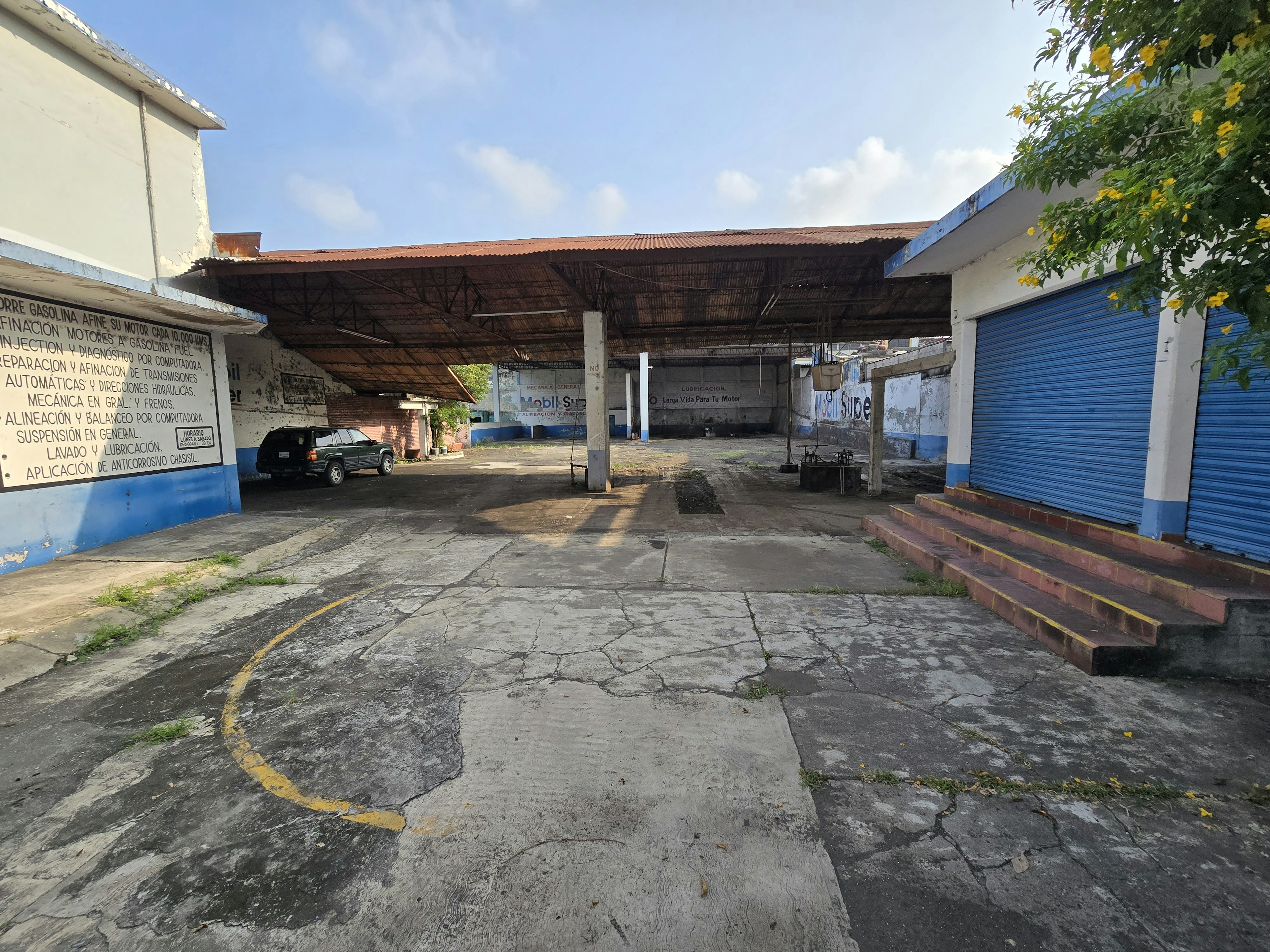 La imagen muestra una estación de servicio o taller mecánico abandonado con un techo de lámina oxidada sostenido por columnas. El piso de concreto está agrietado y tiene marcas amarillas. A los lados se ven edificios blancos con texto en español, uno con detalles azules. Hay vegetación alrededor y un cielo azul claro. El lugar parece estar en desuso y podría ser una oportunidad para remodelación o nuevo desarrollo inmobiliario.