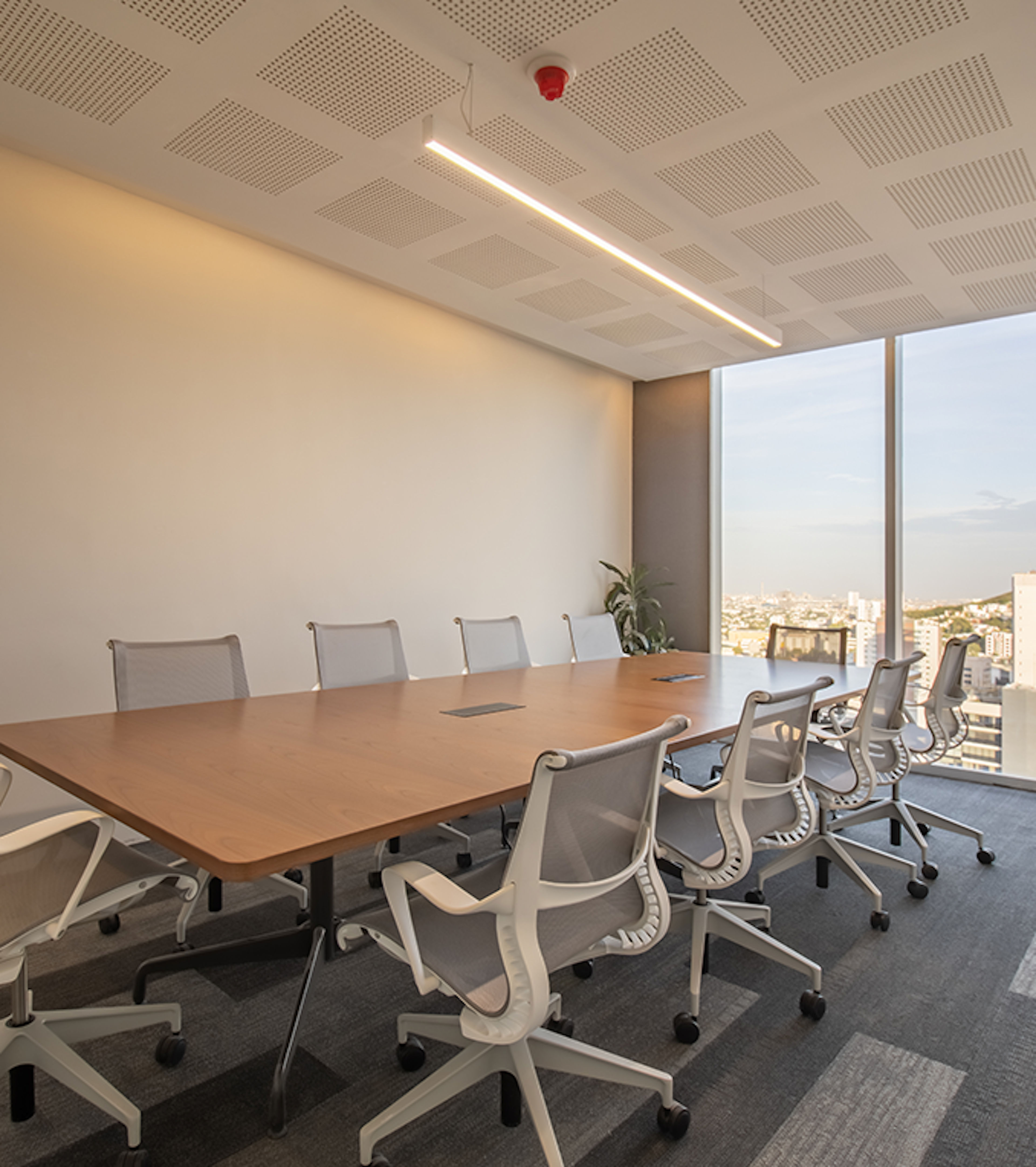 La imagen muestra una moderna sala de reuniones corporativa con una gran mesa de madera y sillas ergonómicas blancas. El techo tiene paneles acústicos y una larga lámpara LED. Una gran ventana ofrece vistas panorámicas de la ciudad. El espacio está diseñado para promover la productividad y el confort, ideal para oficinas de alto nivel en el sector inmobiliario.